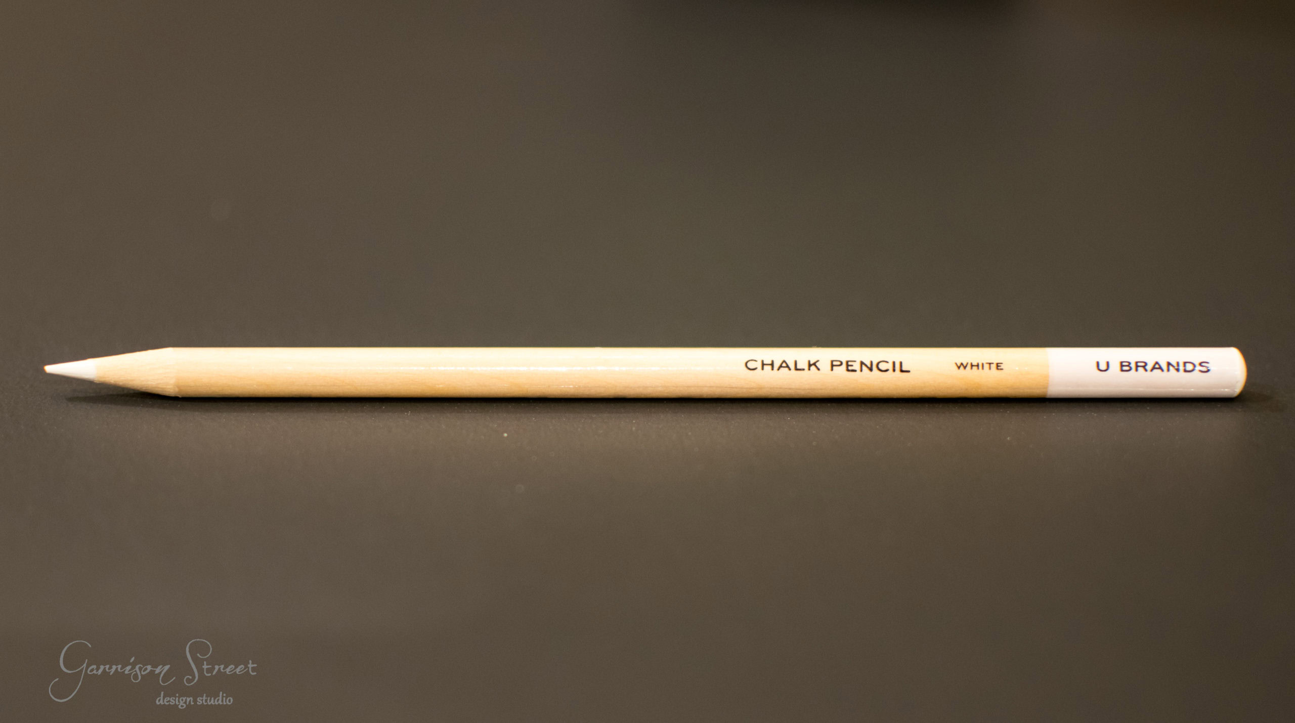 A white chalk pencil with "CHALK PENCIL WHITE" and "U BRANDS" printed on it lies horizontally on a dark surface.