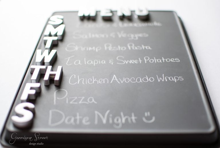 A weekly menu on a chalkboard with magnetic letters for each day. It lists meals: Salmon & Veggies, Shrimp Pesto Pasta, Tilapia & Sweet Potatoes, Chicken Avocado Wraps, Pizza, and Date Night with a smiley face.