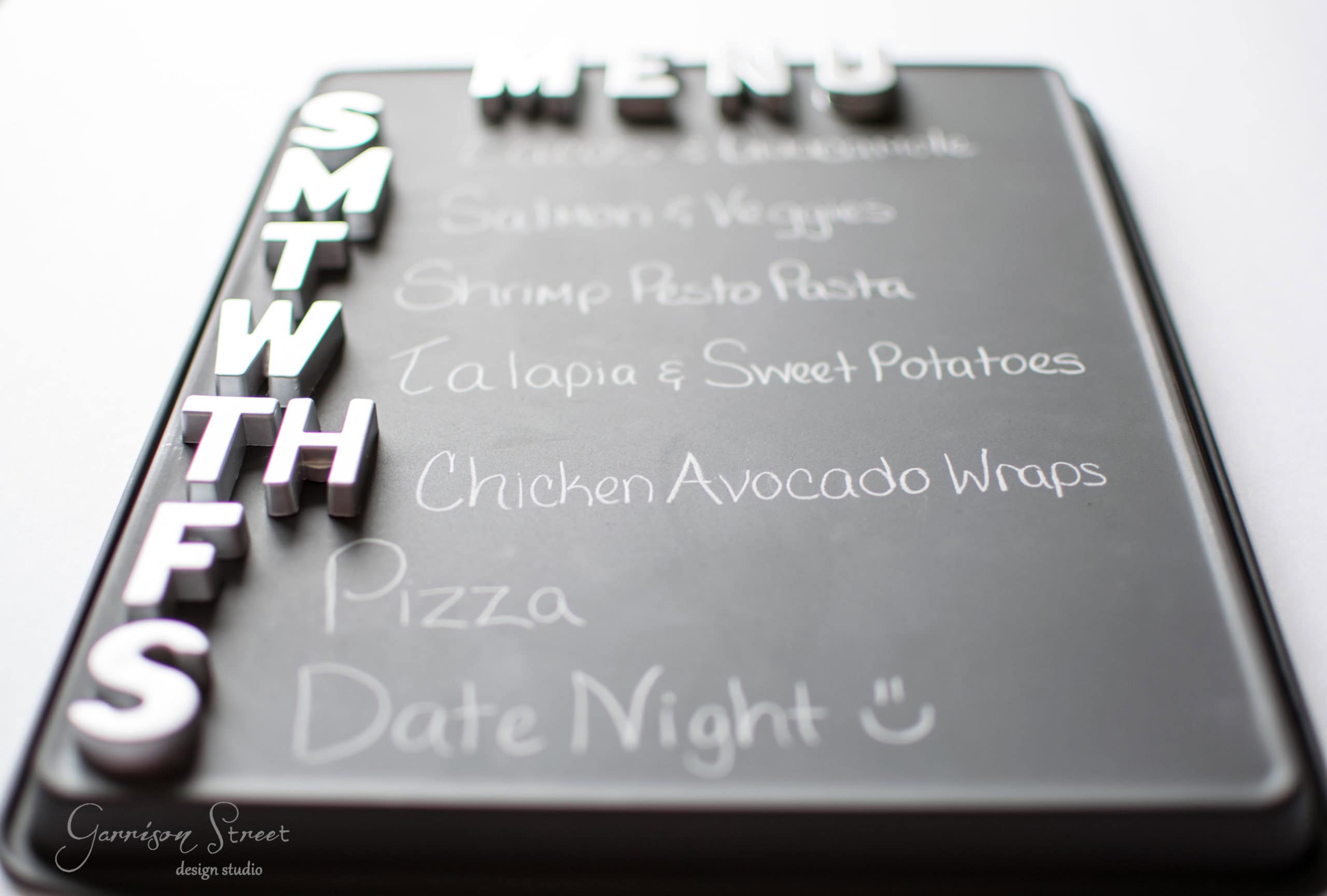 A weekly menu on a chalkboard with magnetic letters for each day. It lists meals: Salmon & Veggies, Shrimp Pesto Pasta, Tilapia & Sweet Potatoes, Chicken Avocado Wraps, Pizza, and Date Night with a smiley face.