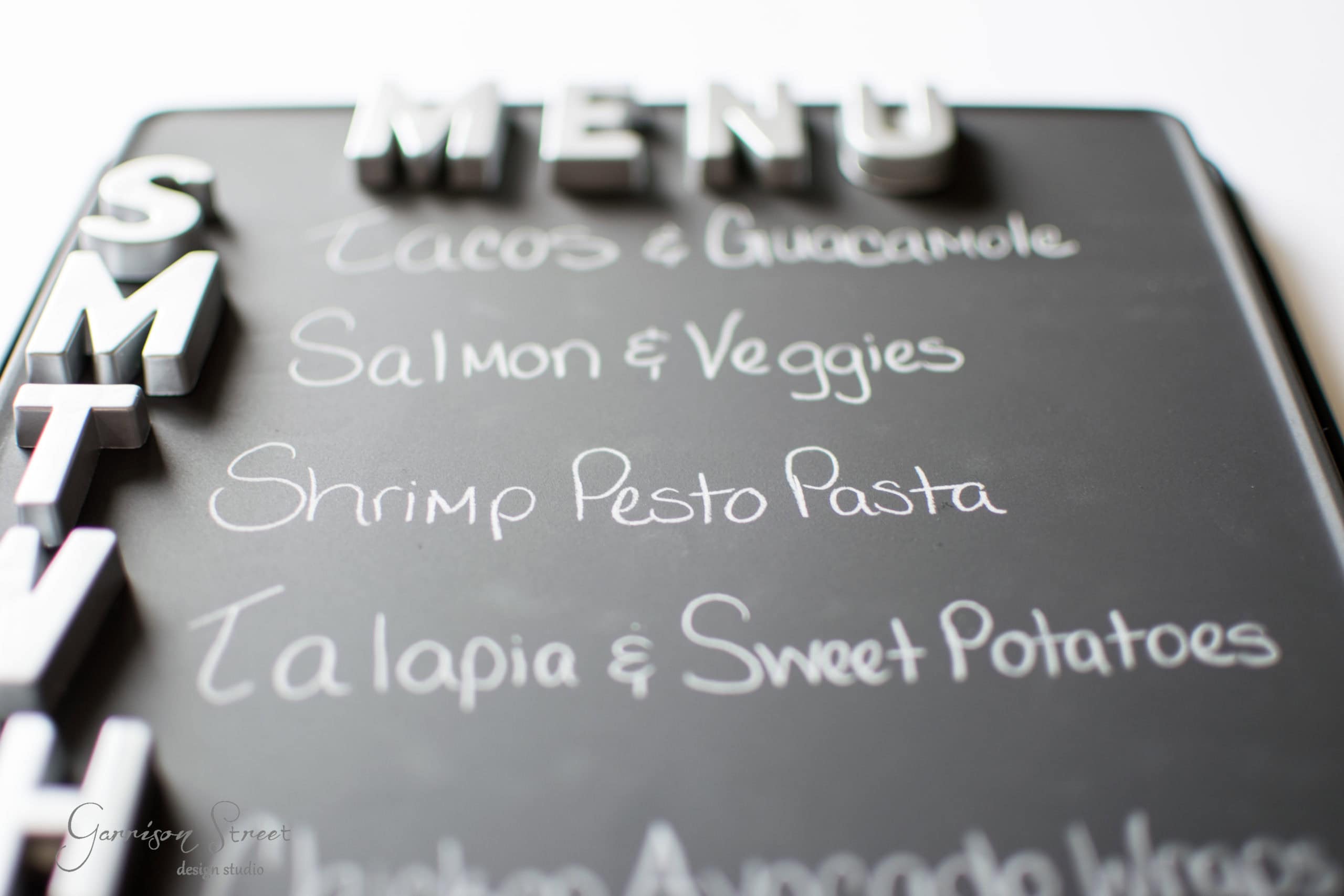 A weekly menu board with letters "SMTWT" vertical on the side. Dishes include Tacos & Guacamole, Salmon & Veggies, Shrimp Pesto Pasta, and Tilapia & Sweet Potatoes. The board also has "MENU" written at the top.