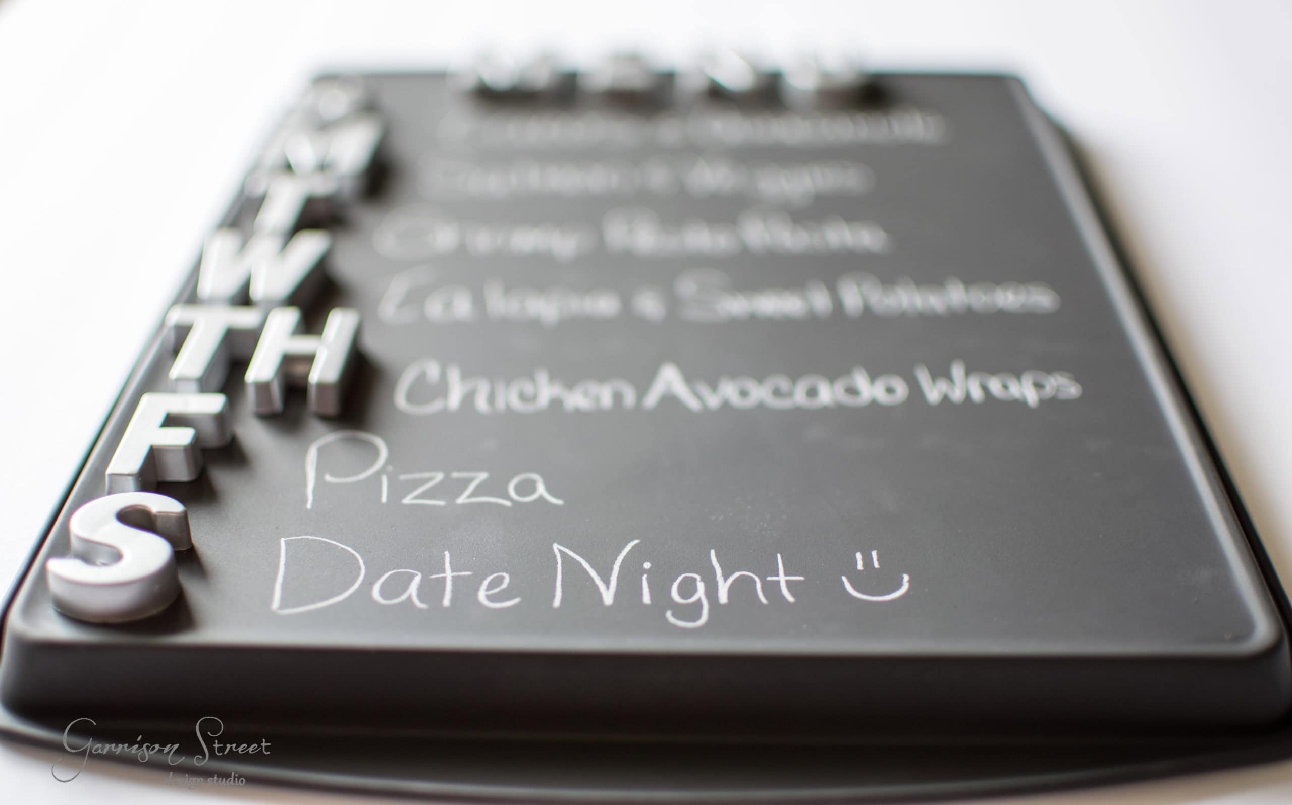 A black chalkboard meal planner with magnetic letters for each day of the week. "Pizza" and "Date Night" are handwritten in chalk for Friday and Saturday. Other days list dishes like "Chicken Avocado Wraps.