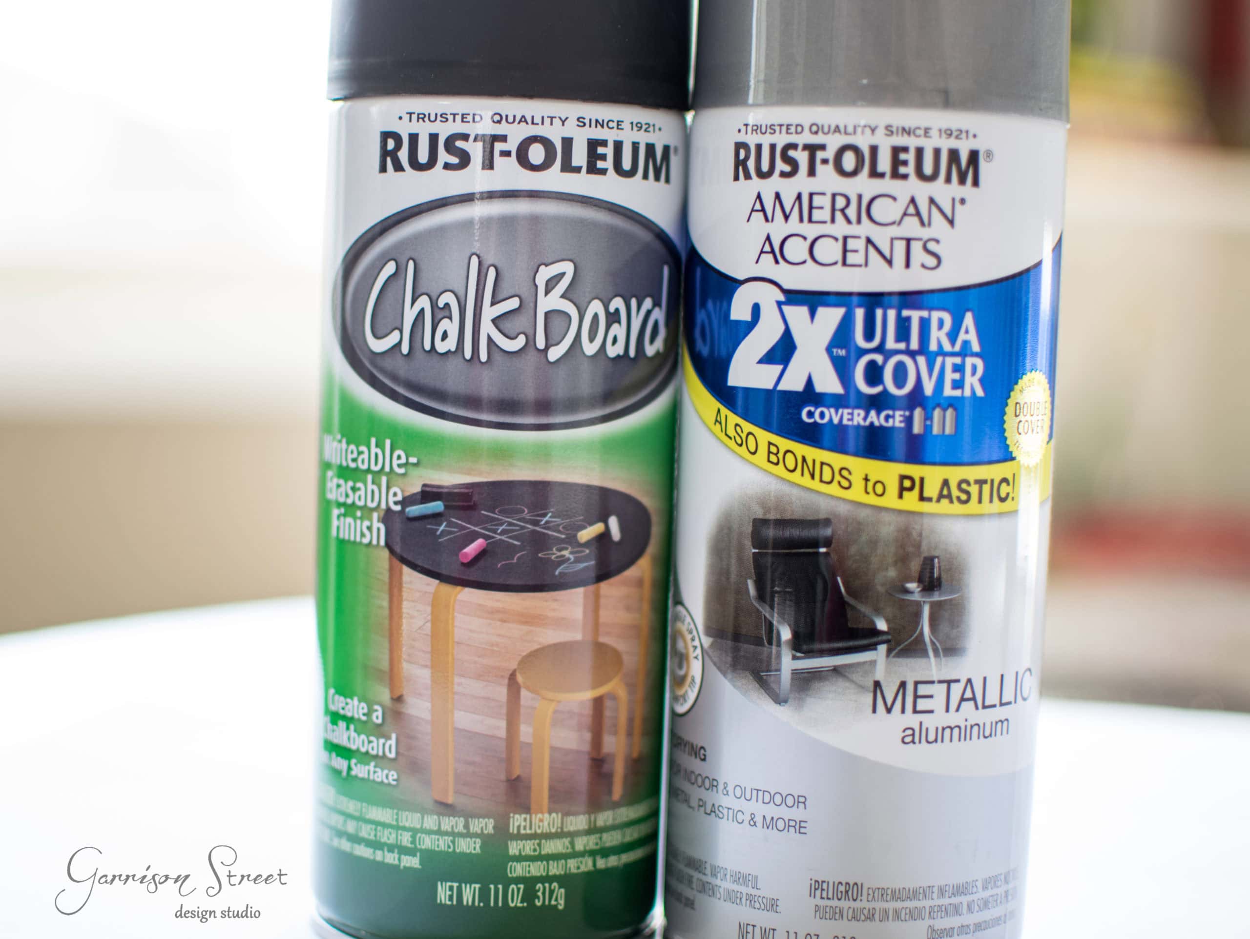 Close-up of two spray paint cans. Left can: Rust-Oleum Chalkboard spray for writable surfaces, featuring a table graphic. Right can: Rust-Oleum American Accents 2X Ultra Cover in metallic aluminum finish, with "Also Bonds to Plastic!" text.