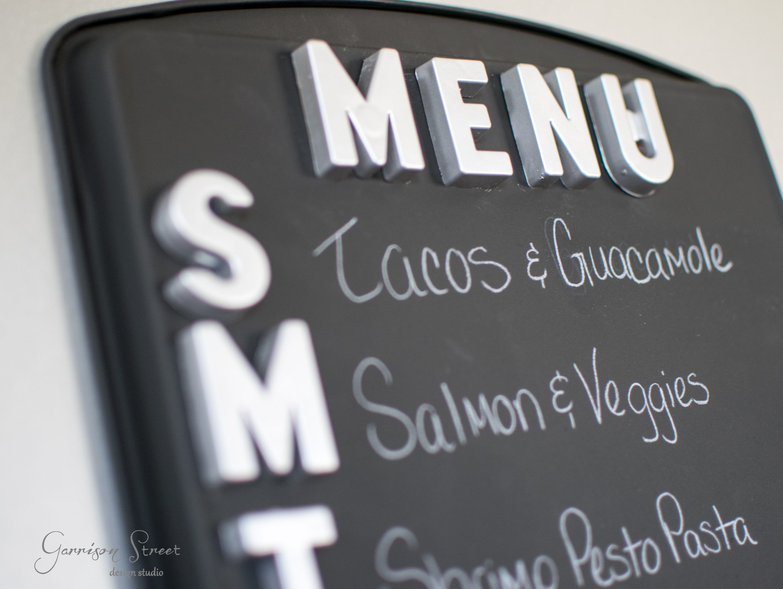 Close-up of a menu board with 3D letters. The days are abbreviated on the left: S, M, T. Dishes listed include Tacos & Guacamole, Salmon & Veggies, and Shrimp Pesto Pasta.