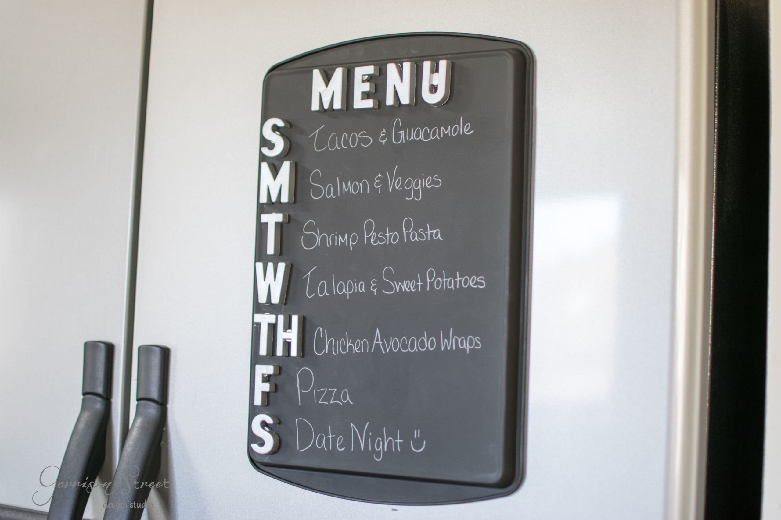 A magnetic menu board on a fridge lists meals for each day: tacos and guacamole, salmon and veggies, shrimp pesto pasta, tilapia and sweet potatoes, chicken avocado wraps, pizza, and date night. Days of the week are labeled from Sunday to Saturday.