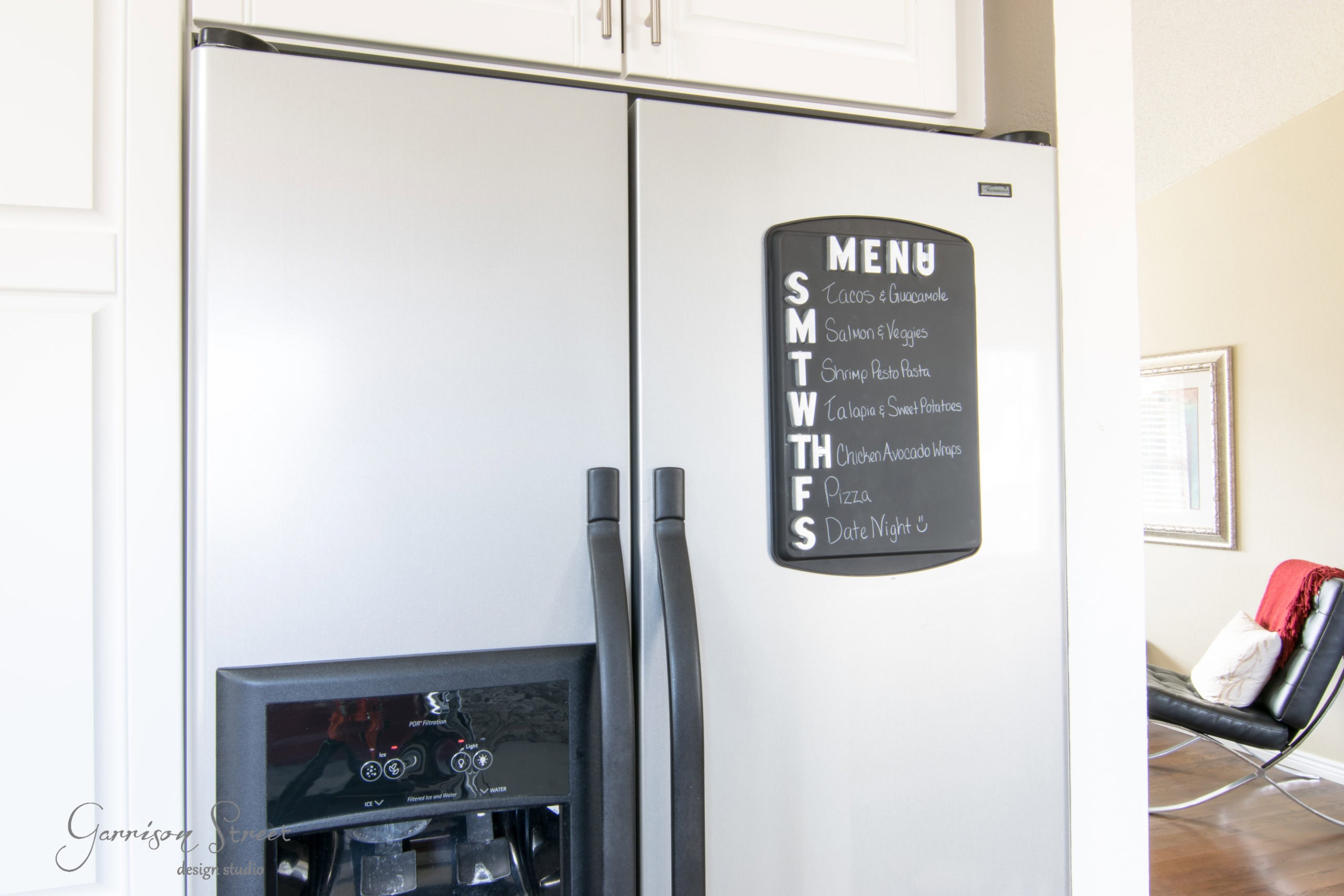 A stainless steel refrigerator with a chalkboard menu on the door. The menu lists meals for each day of the week, from Sunday to Saturday. A chair and part of the room are visible in the background.