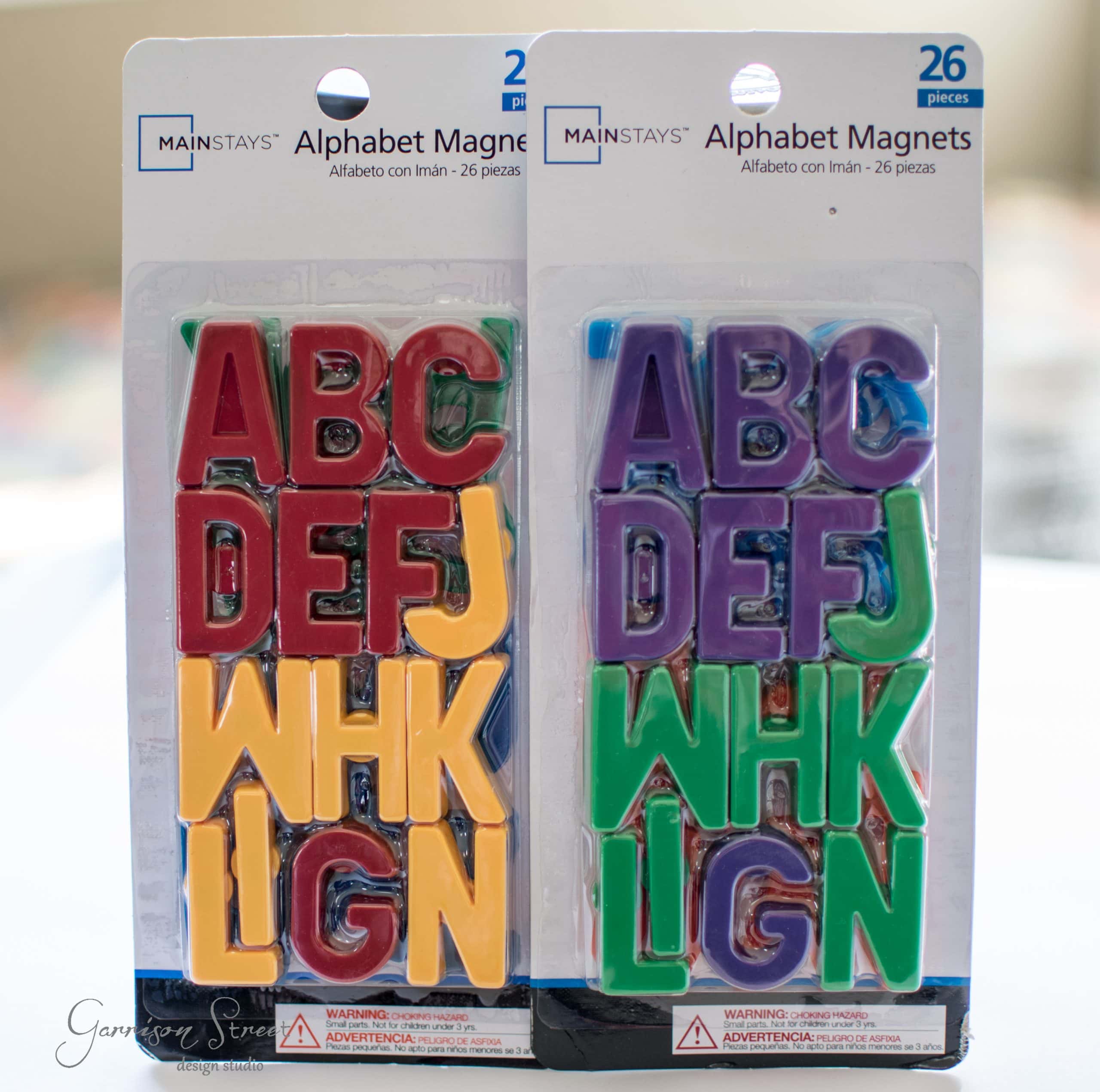 Two packages of colorful alphabet magnets are displayed. Each package contains 26 pieces with letters in red, yellow, blue, green, and purple. The packaging is labeled "Alphabet Magnets" with warnings in English and Spanish.