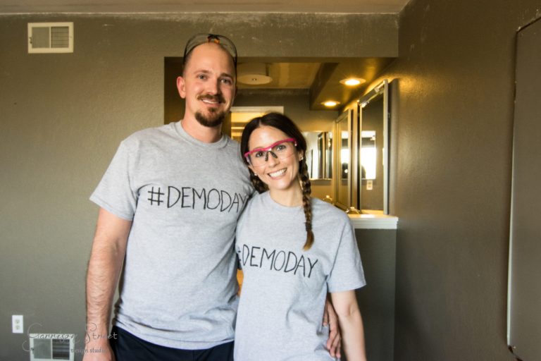 A smiling couple stands indoors wearing matching gray t-shirts with "#DEMODAY" printed on them. The woman wears pink safety glasses and has her hair in a braid. They appear to be in a lightly renovated room.