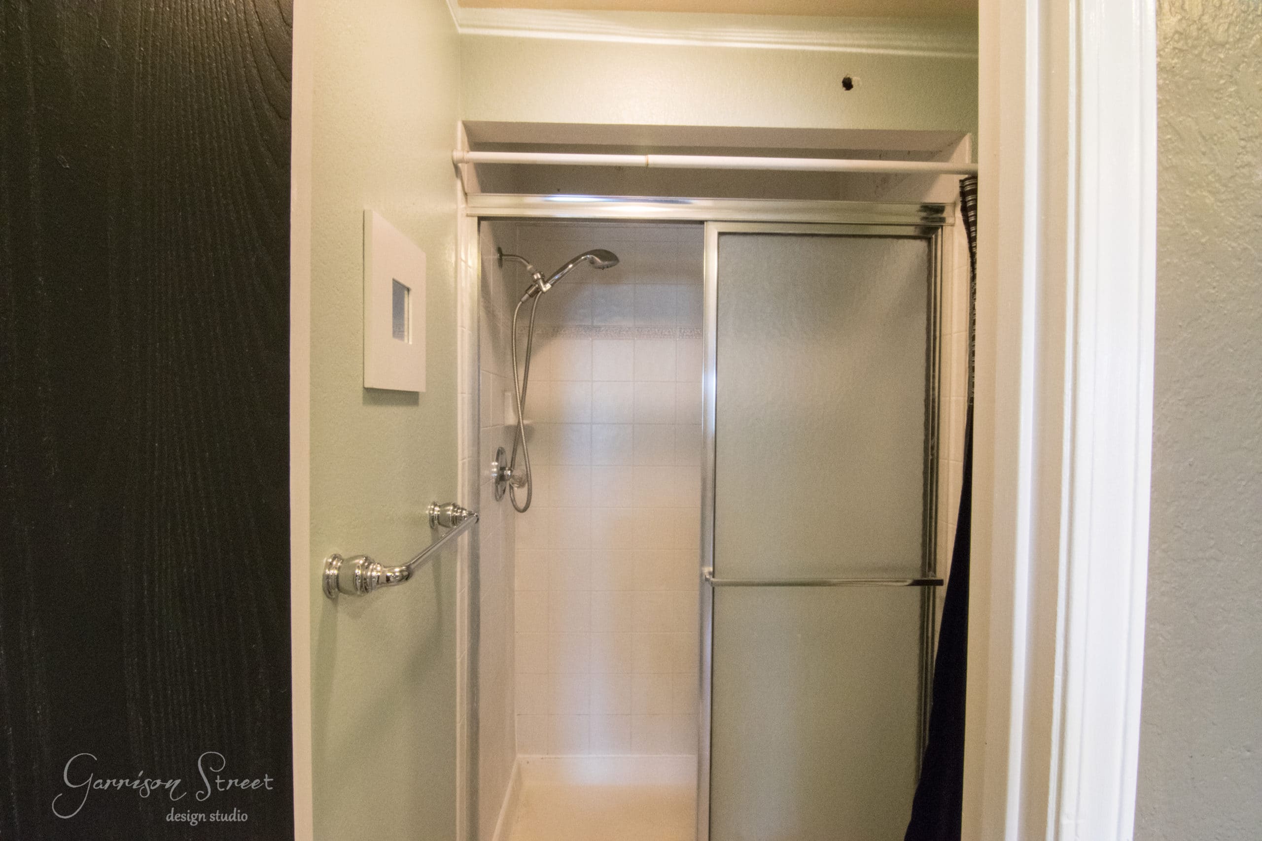 A small bathroom with a shower area featuring sliding glass doors. The shower has white tiles and a wall-mounted showerhead. The walls are painted in a light green color, and a towel rack is mounted on the side wall.