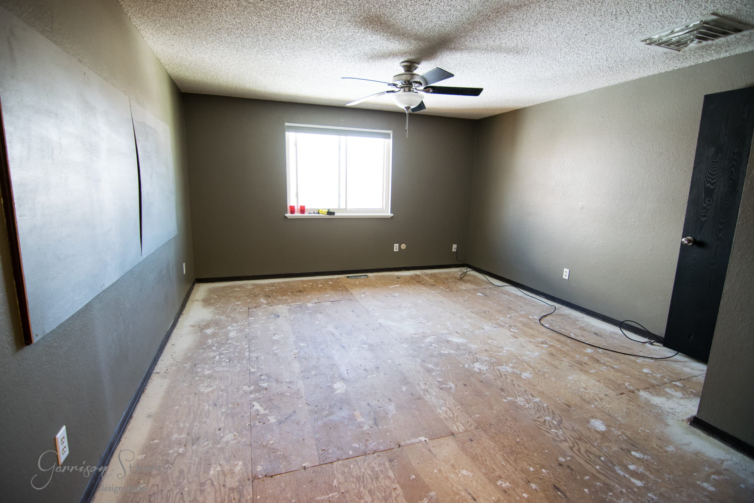 An empty room with exposed plywood flooring and gray walls. A ceiling fan is centered above, and a single window allows natural light. A black door is on the right, and a few electrical outlets are visible along the walls.