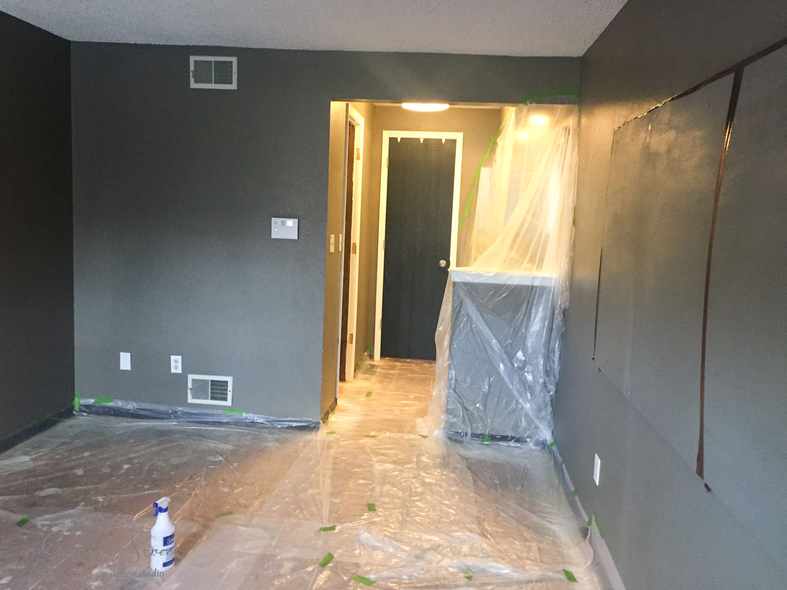 A room with dark painted walls and a floor covered in plastic sheeting, leading to a hallway. Green tape secures the plastic. A spray bottle is on the floor, and a plastic sheet covers part of a cabinet in the hallway.