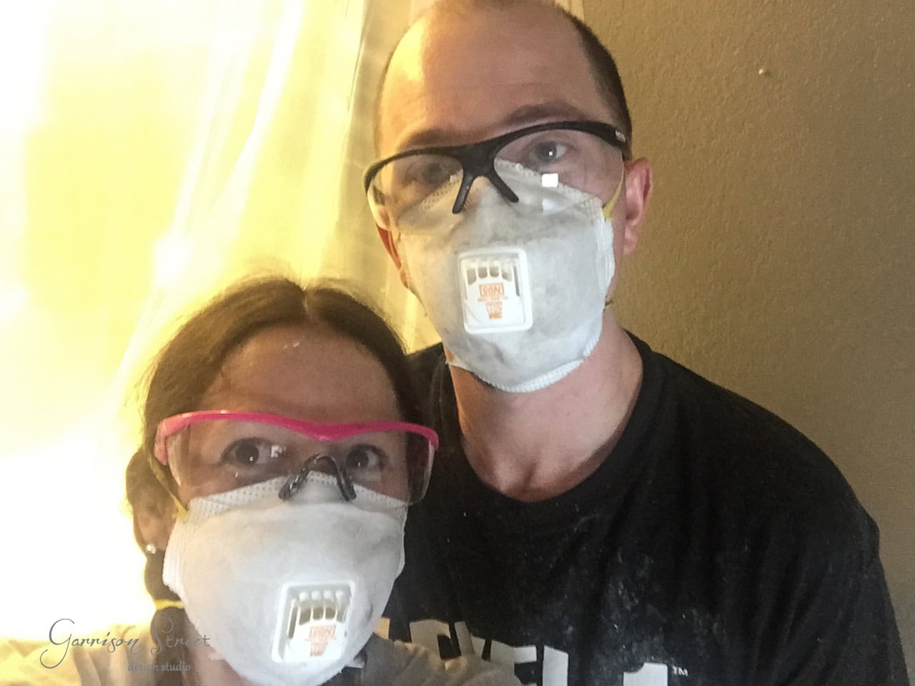 Two people wearing protective masks and goggles stand close together. The person on the left has bright pink goggles, while the person on the right has black goggles. They are indoors, with a curtain in the background.