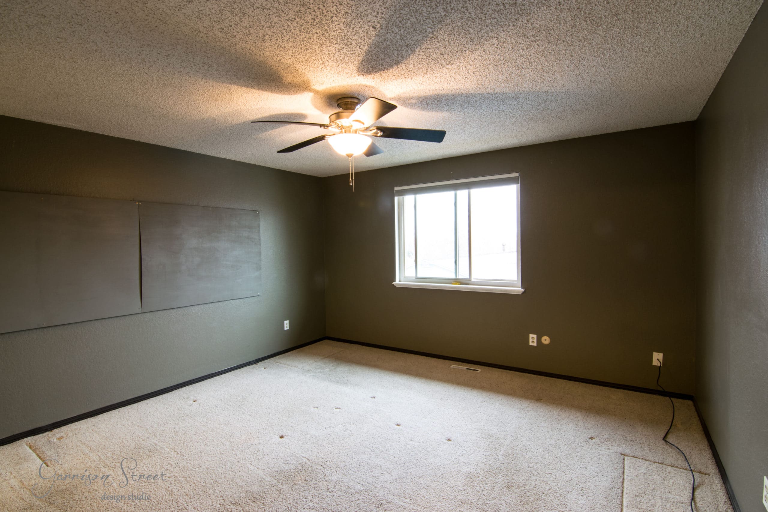 An empty room with dark green walls, a ceiling fan with a light, and a window allowing natural light. The floor is carpeted, and a large black board or panel is mounted on one wall.