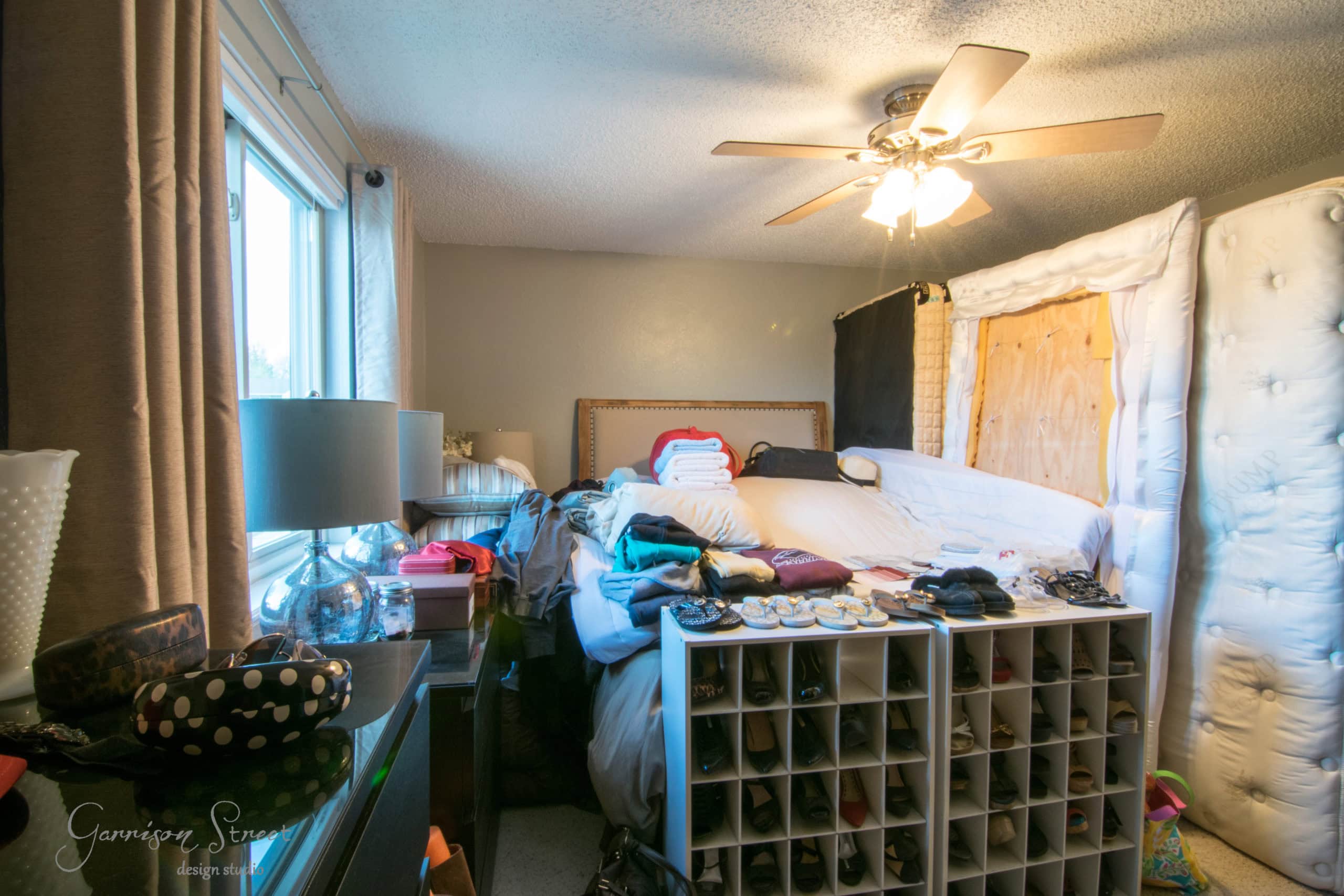 A cluttered bedroom with a bed piled high with clothes and a shoe organizer filled with shoes. A lamp sits on a dresser near the window, and a ceiling fan is on a beige ceiling. A white headboard and a curtain are visible.