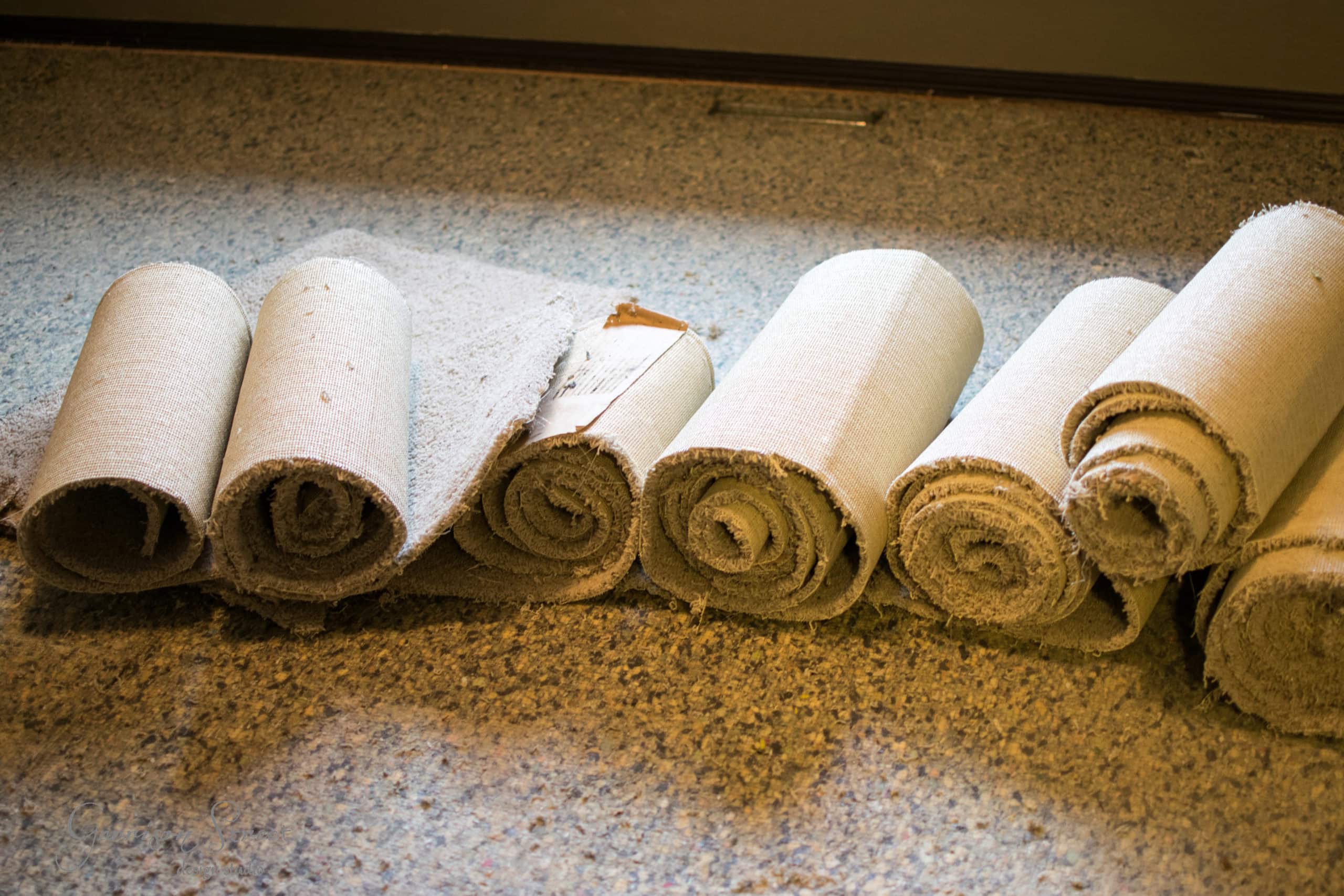 Rolled-up pieces of old carpet lined up on a speckled carpeted floor, showing signs of wear and tear.