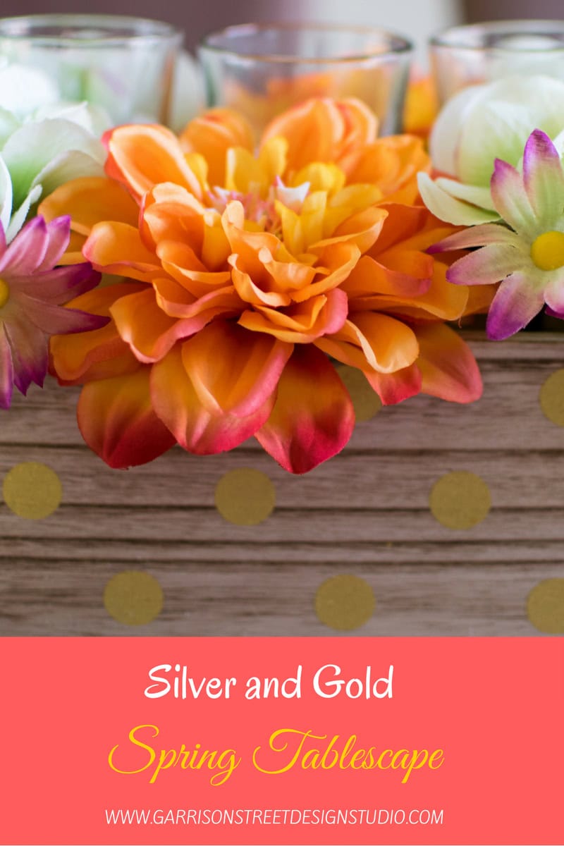 A close-up of a vibrant spring tablescape with an arrangement of orange and pink flowers in front of votive candles. The background features a wooden board with gold polka dots. Text reads: "Silver and Gold Spring Tablescape.