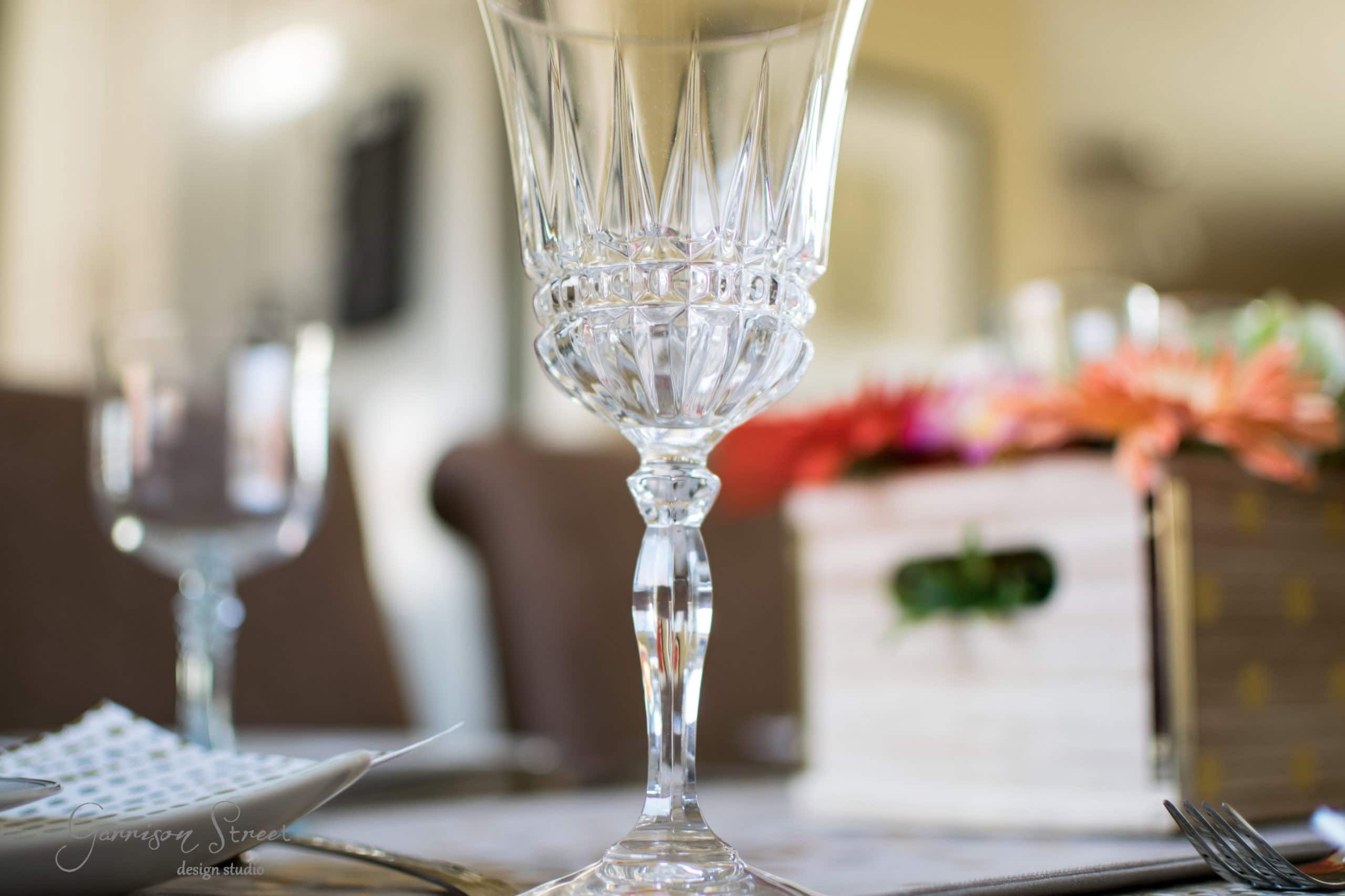 A close-up of an elegant crystal wine glass situated on a dining table. In the background, there's a wooden box with colorful flowers and blurred tableware, creating a sophisticated and inviting atmosphere.