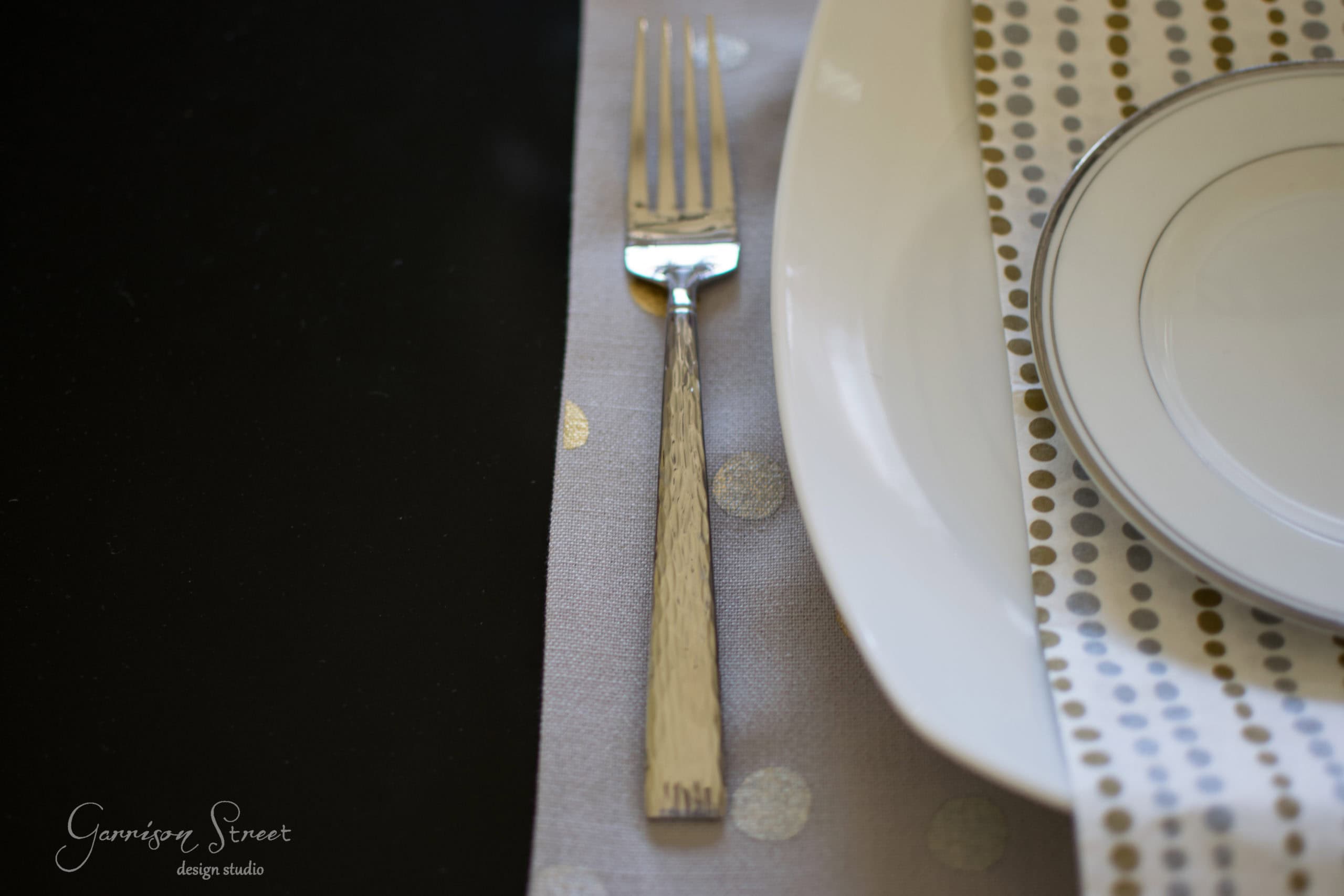 A neatly arranged table setting with a silver fork on a gray and gold polka-dot napkin beside an elegant white plate with a gold-rimmed edge. A patterned tablecloth adds a touch of sophistication.