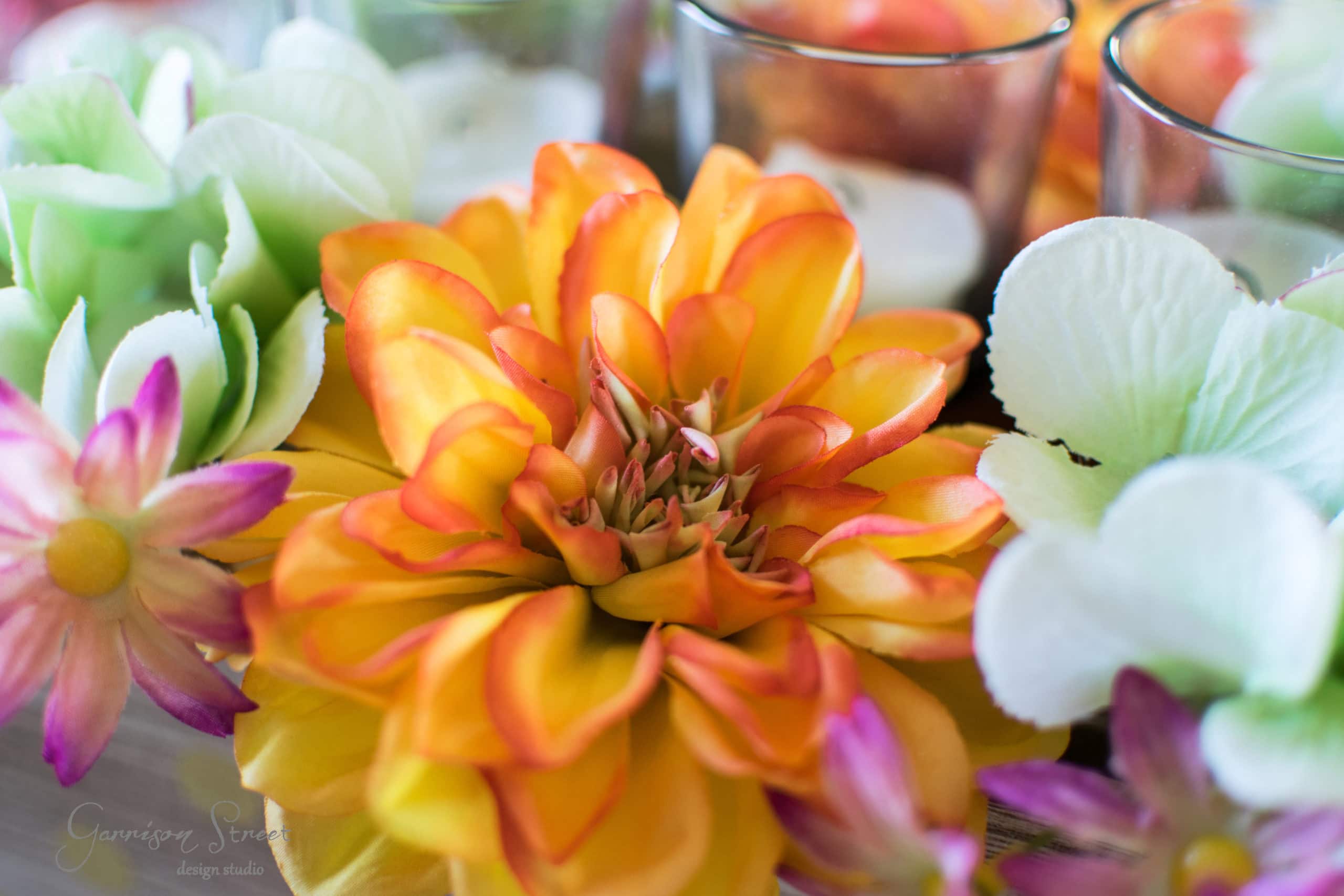 A vibrant close-up of colorful artificial flowers, including a central yellow and orange bloom, surrounded by soft green and white hydrangea petals, with glass candle holders in the background.