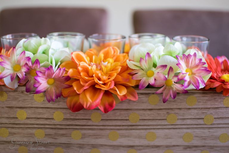 A wooden box with gold polka dots holds vibrant artificial flowers, featuring large orange blooms and smaller pink and white flowers. Glass candle holders are nestled among the flowers in the background.