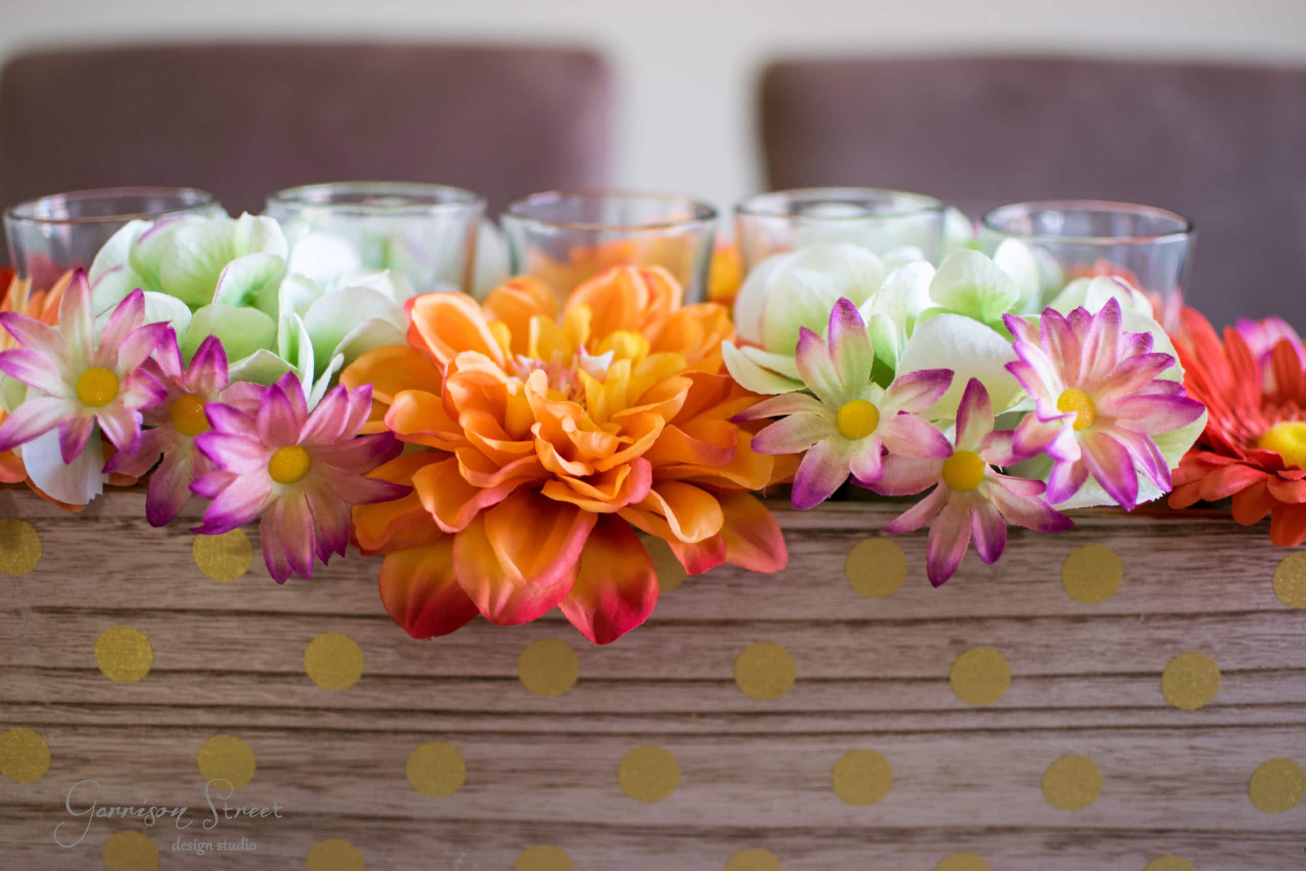 A wooden box with gold polka dots holds vibrant artificial flowers, featuring large orange blooms and smaller pink and white flowers. Glass candle holders are nestled among the flowers in the background.