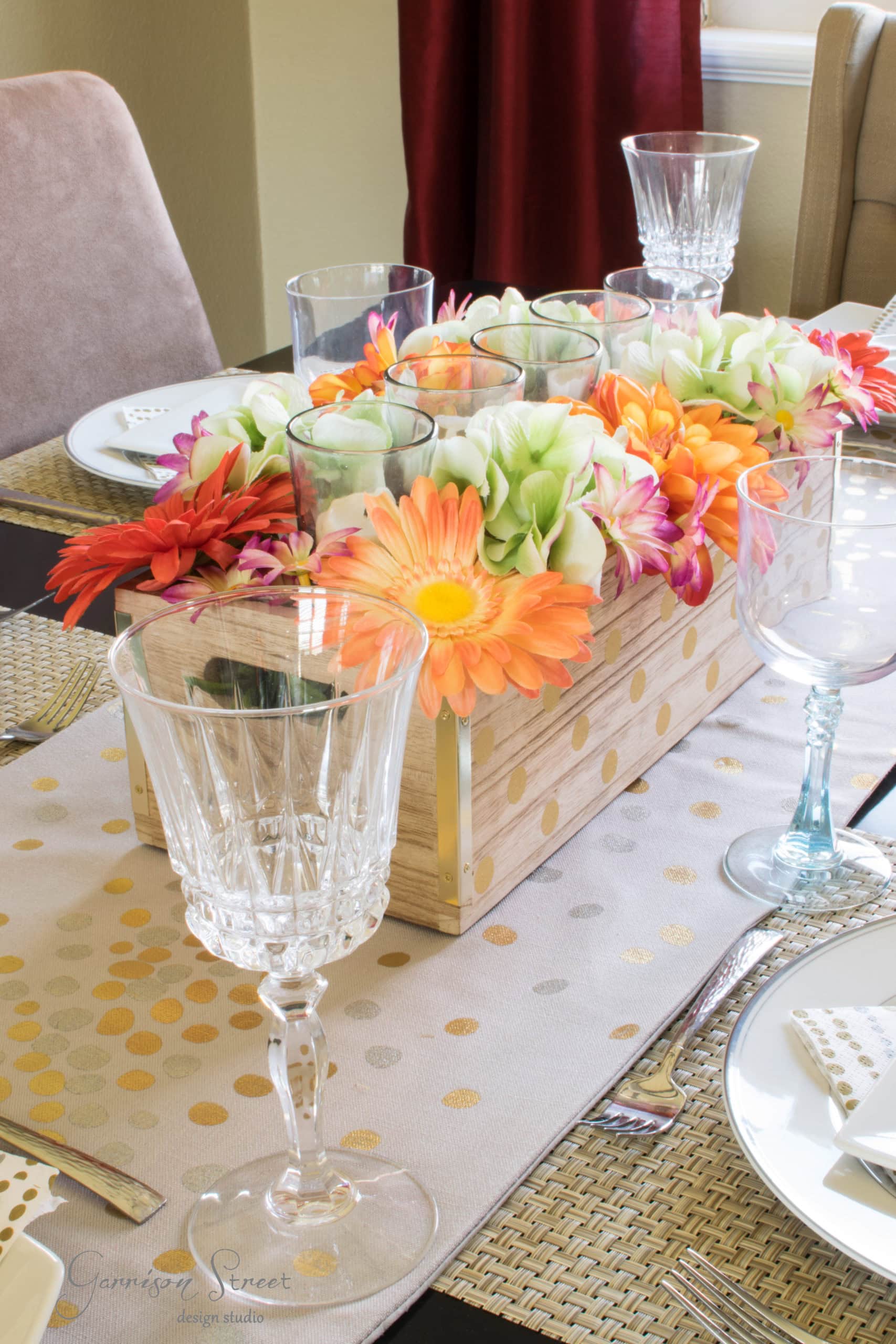 A dining table set with a floral centerpiece featuring vibrant orange, pink, and white flowers in a wooden vase. Crystal wine glasses and plates with silverware are arranged on a decorative table runner with gold dots.
