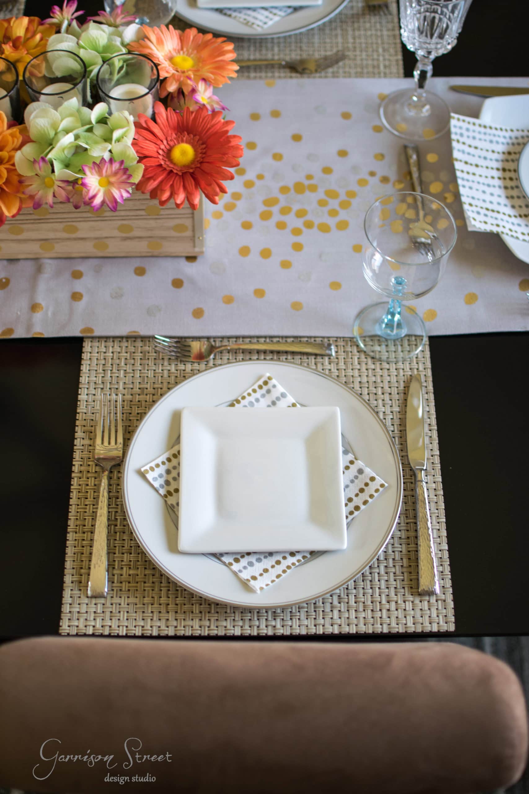 A dining table set with a woven placemat, square white plates, and a dotted napkin. Beside the plates are a glass and cutlery. A centerpiece with colorful flowers and a table runner with gold dots completes the arrangement.
