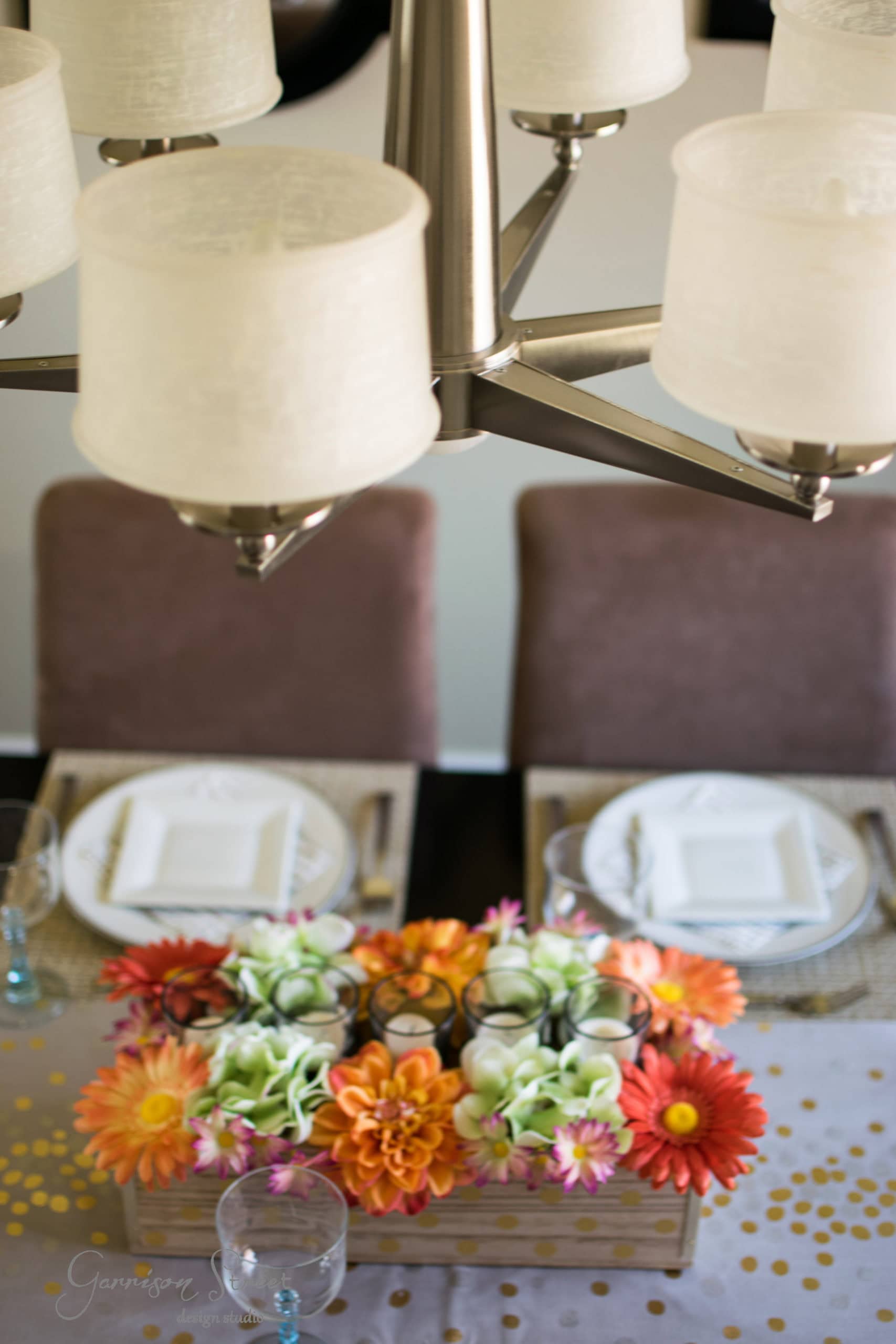 A dining table set for two with white plates and glasses on woven placemats. The centerpiece features an arrangement of colorful flowers, including orange and pink blooms. A chandelier with cream lampshades hangs above the table.
