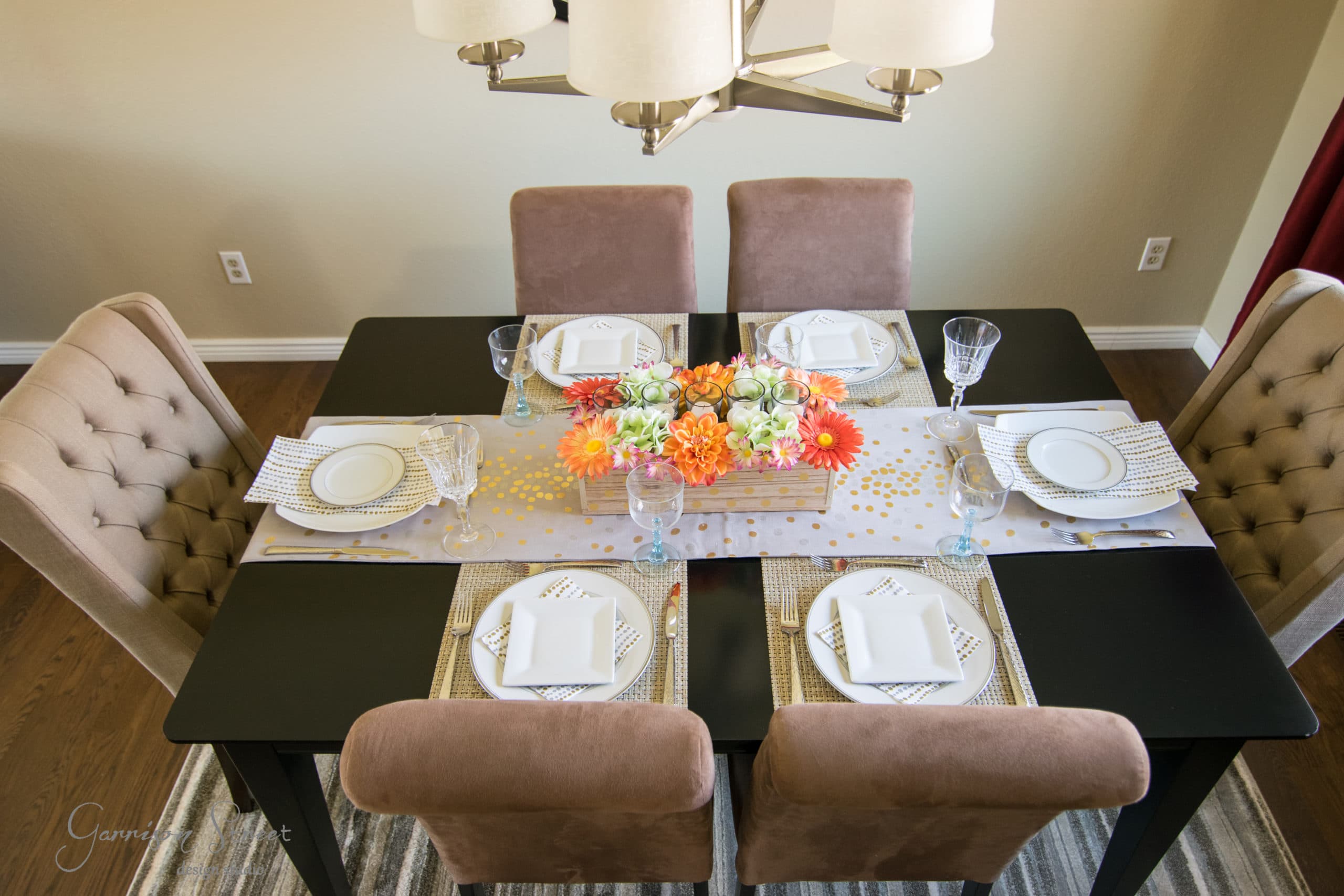 A dining table set for six with beige chairs, featuring white plates and glasses on a black table. The centerpiece is a wooden box filled with orange and yellow flowers. A striped rug is below the table.