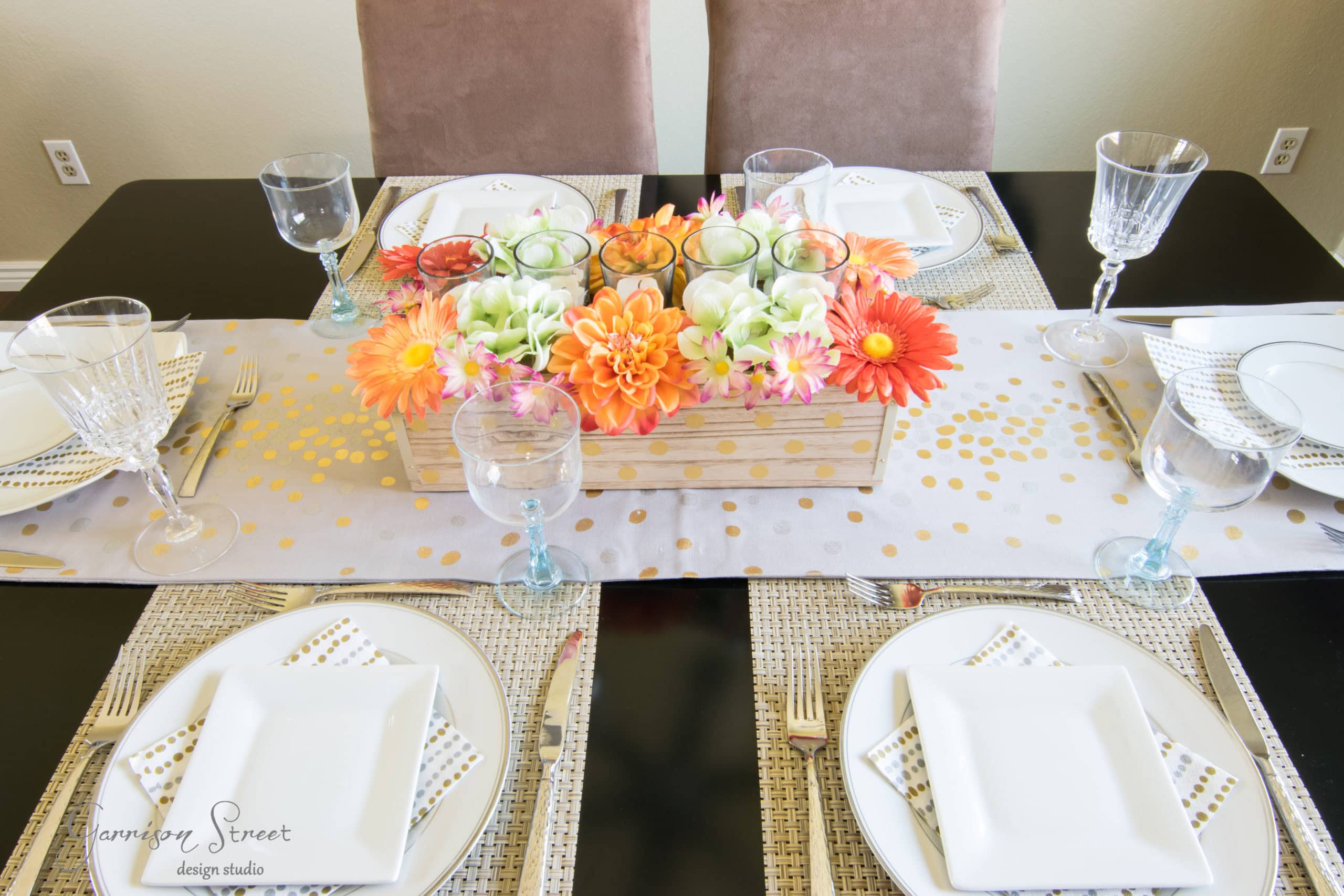 A dining table set for four with white square plates, clear wine glasses, and blue water goblets. The centerpiece is a wooden box with orange and pink flowers arranged around three candles. The table runner is white with gold dots.