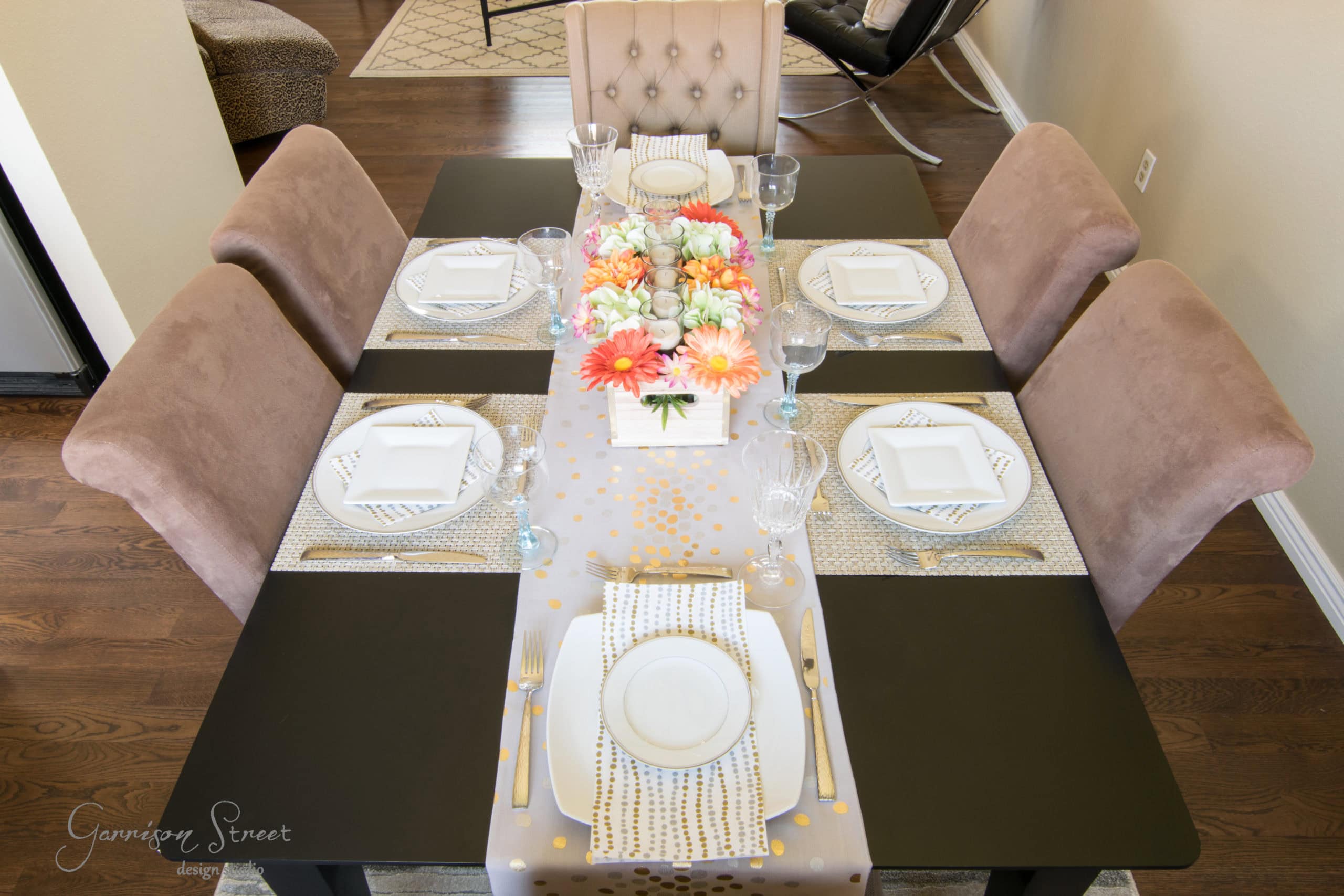 A dining table set for six with a floral centerpiece featuring orange and white flowers. The table has white plates, glasses, and striped napkins on beige placemats. The chairs are upholstered in light brown fabric, and the room has wooden flooring.