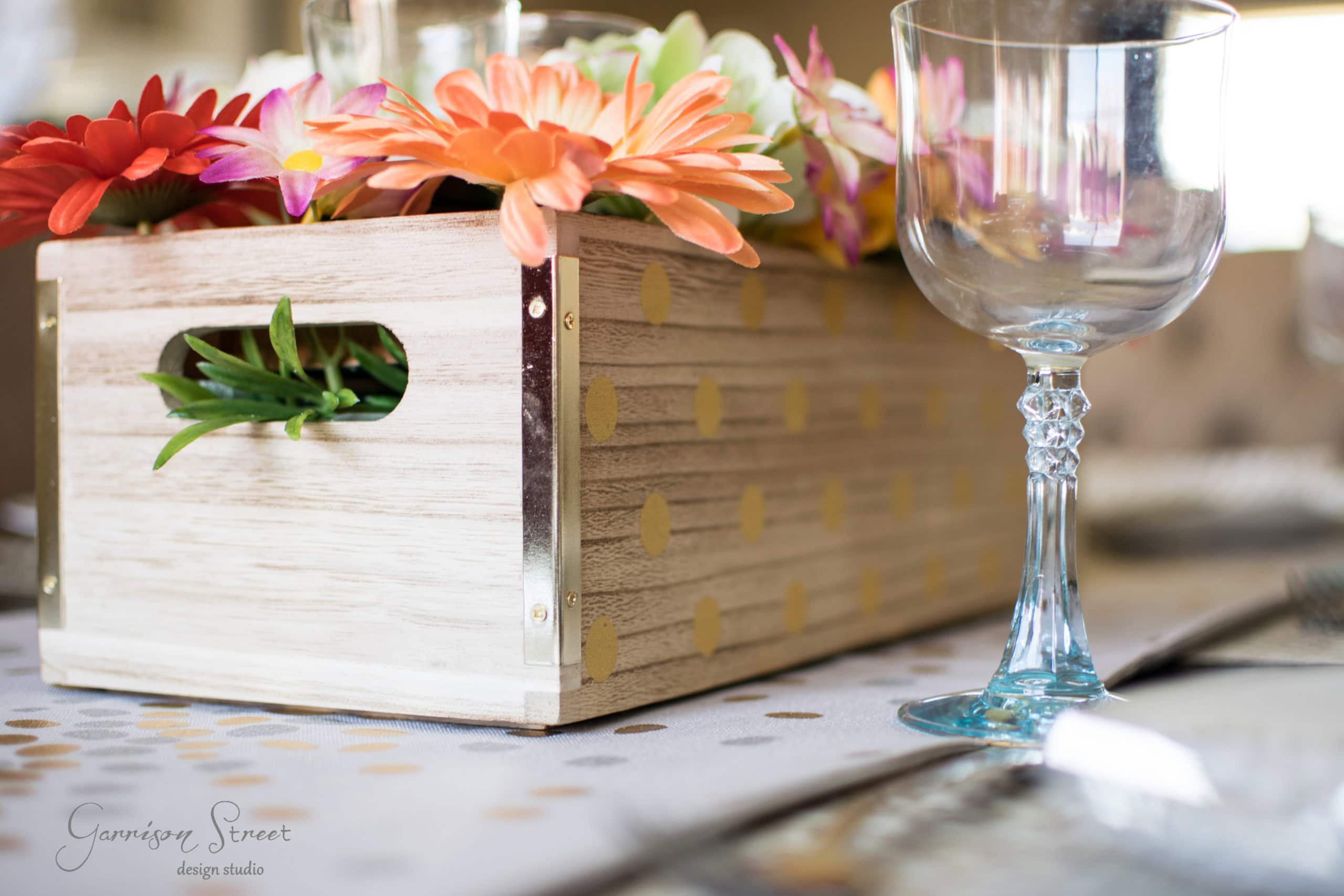 Wooden box with colorful flowers sits on a table next to an empty wine glass. The box has gold polka dots, and the tablecloth features a soft polka dot pattern. The setting is bright and elegant.