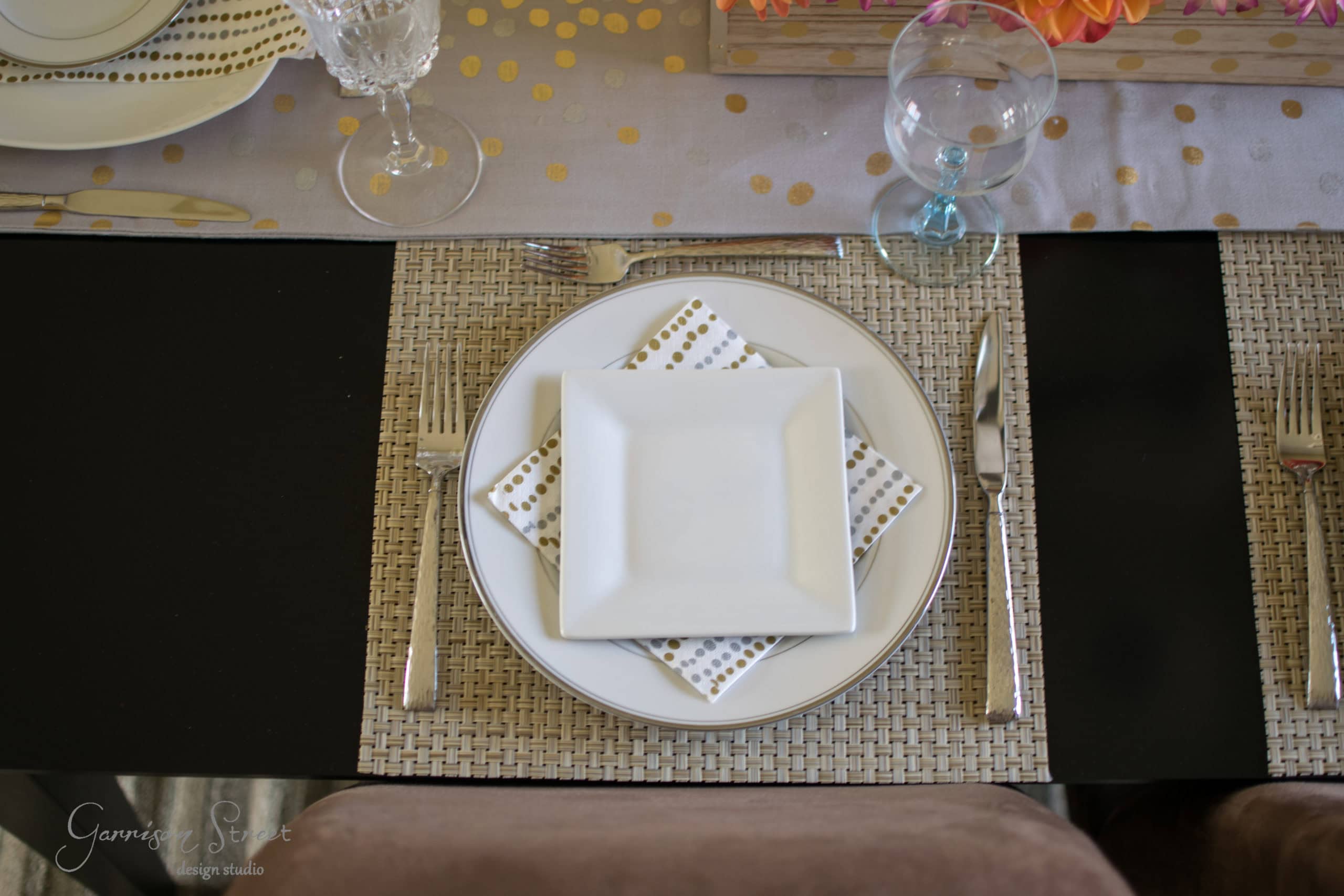 A neatly arranged place setting on a woven placemat features a square white plate on a dotted napkin, surrounded by silverware. A glass of water and a wine glass are placed above, with a table runner and orange flowers in the background.