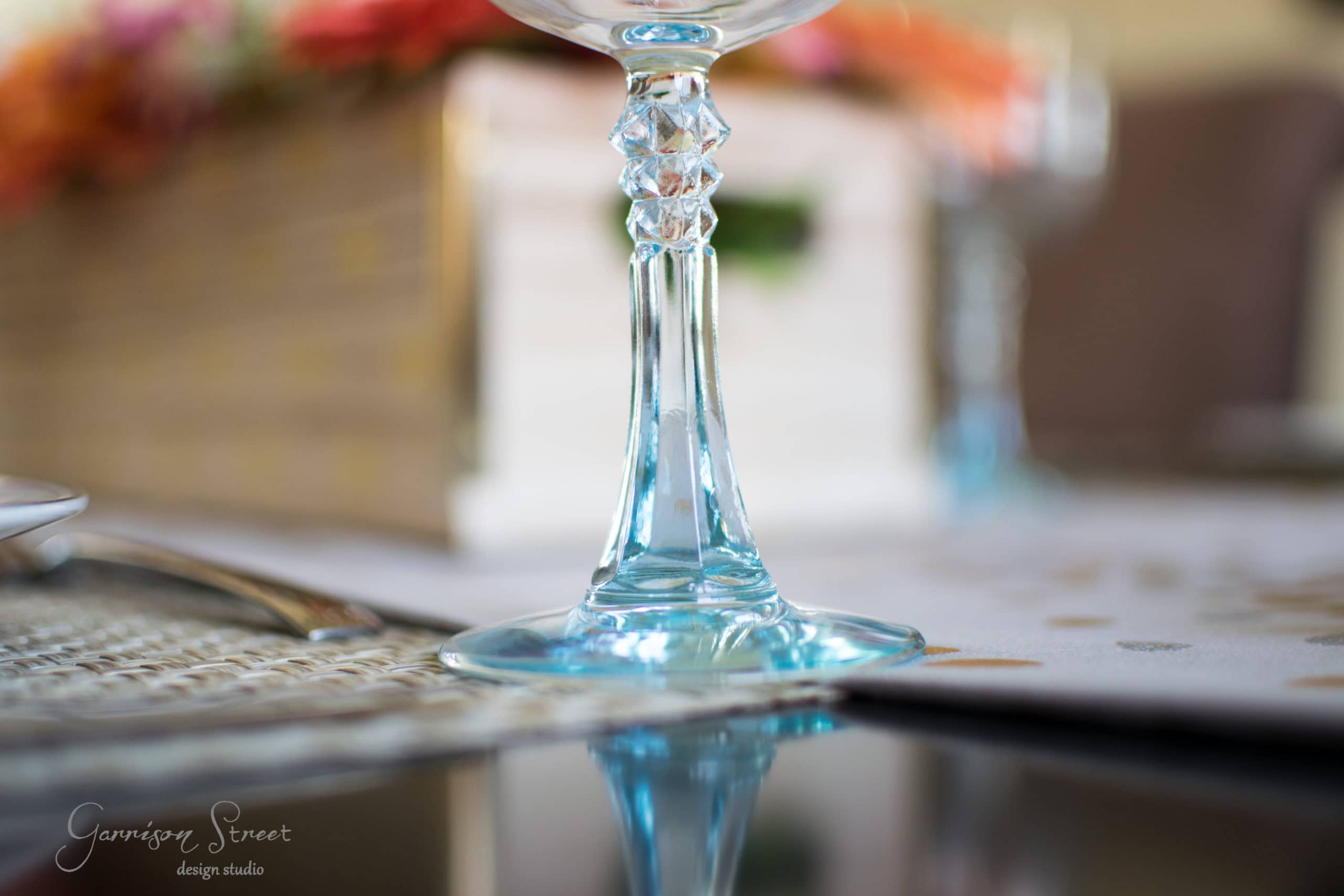 Close-up of a crystal wine glass stem on a reflective surface, with blurred pastel flowers in the background. The image captures an elegant table setting with soft lighting and a focus on details.