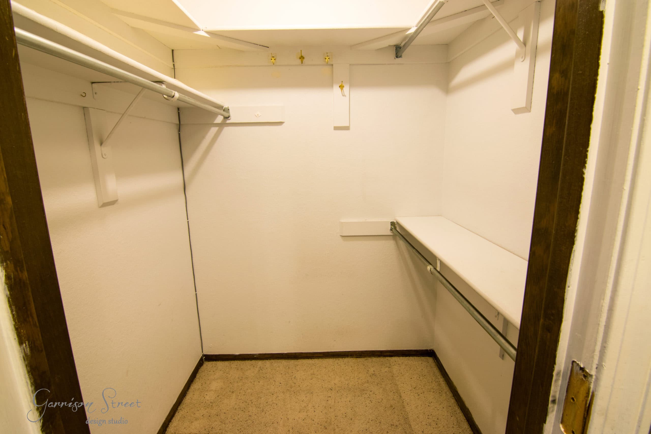 A small, empty walk-in closet with white walls and wooden trim. It features a shelf and rod on the right side, additional hooks on the walls, and a bare floor. The ceiling appears slightly slanted.