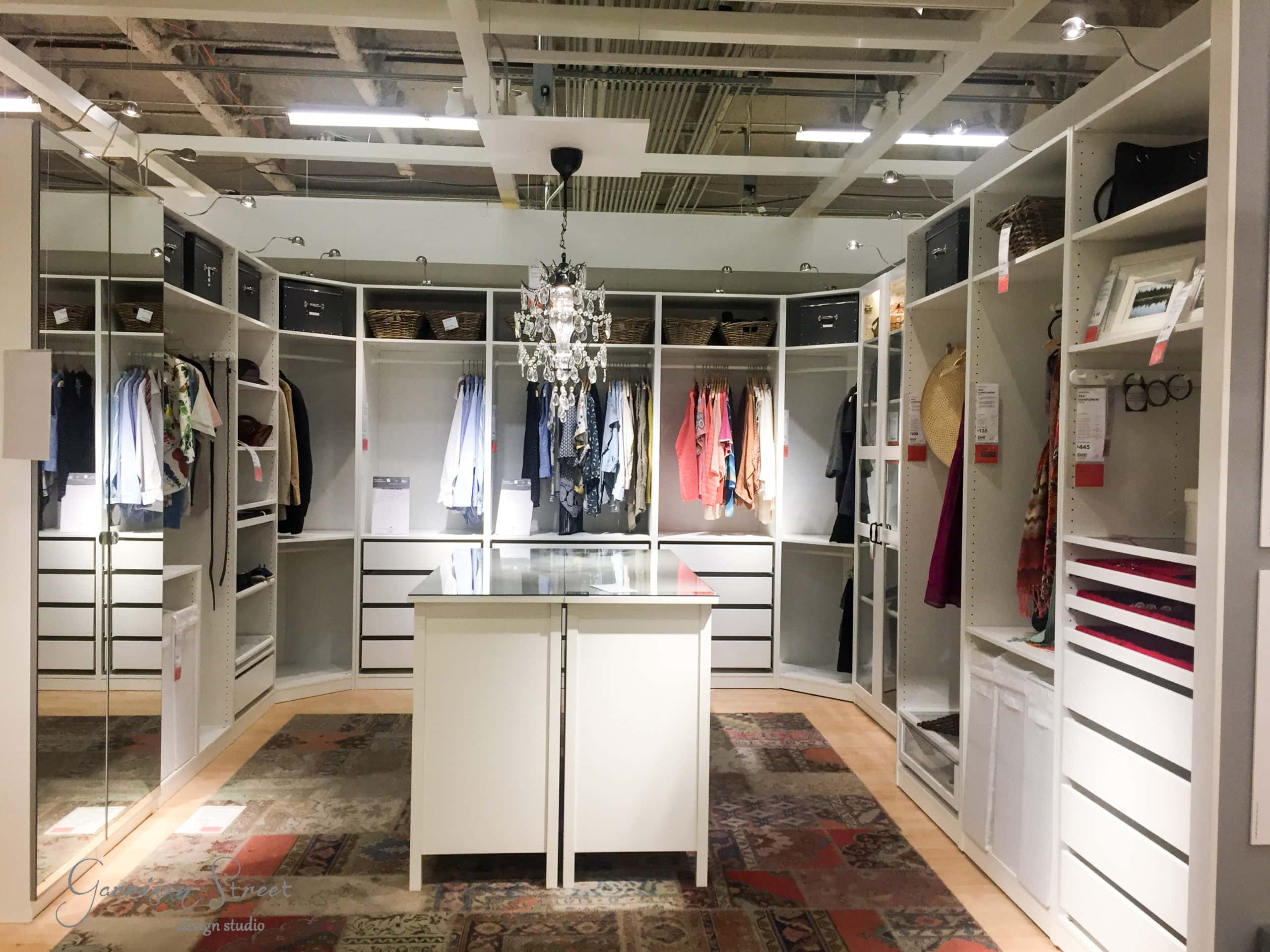 A spacious walk-in closet with neatly arranged clothes, shoes, and accessories on white shelves. A central island with drawers sits beneath a chandelier. Warm lighting highlights the room's organized, modern design.
