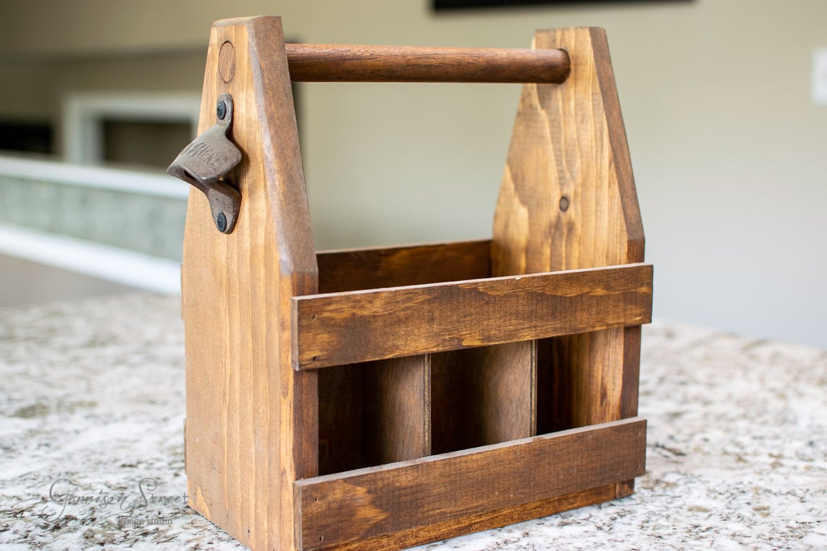 A wooden condiment caddy with a handle, featuring an attached bottle opener, rests on a marble countertop. The caddy has a rustic finish and is perfect for holding bottles or other items.