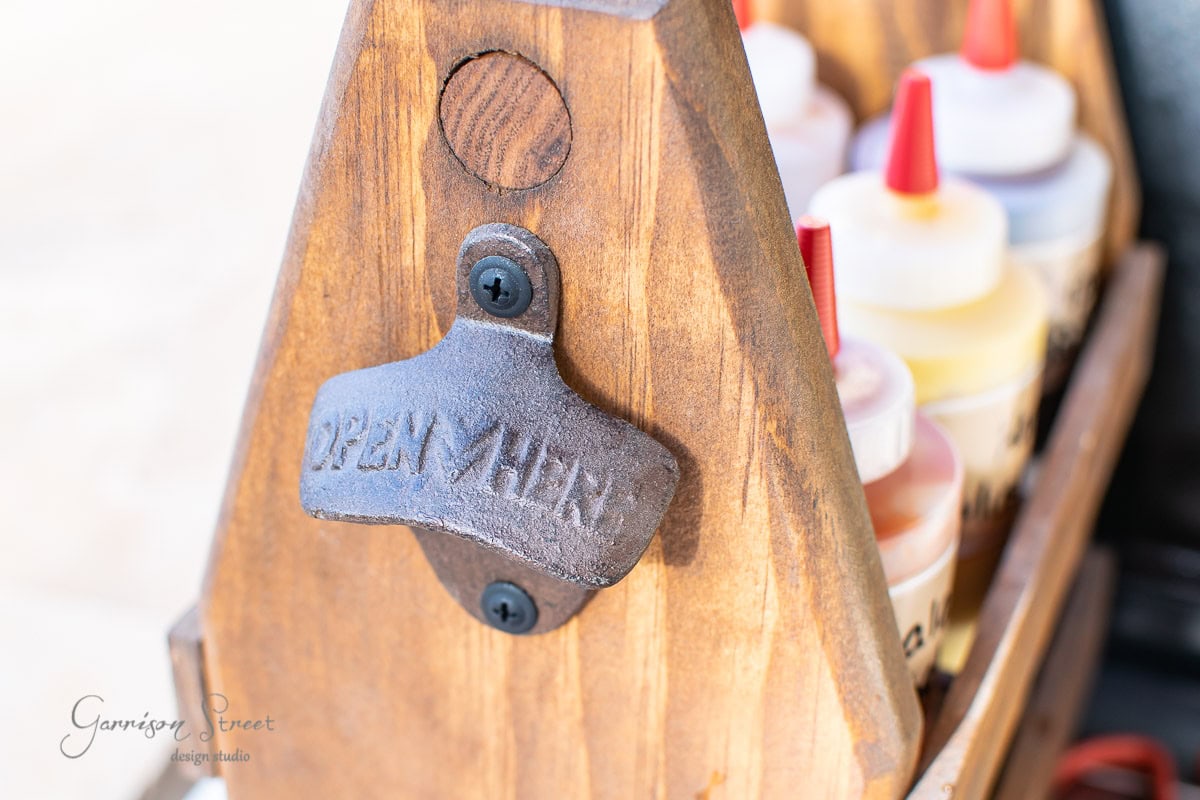 A wooden condiment caddy with a black metal bottle opener labeled "Open Here." Inside the caddy, several small plastic bottles with red and yellow caps are neatly arranged.