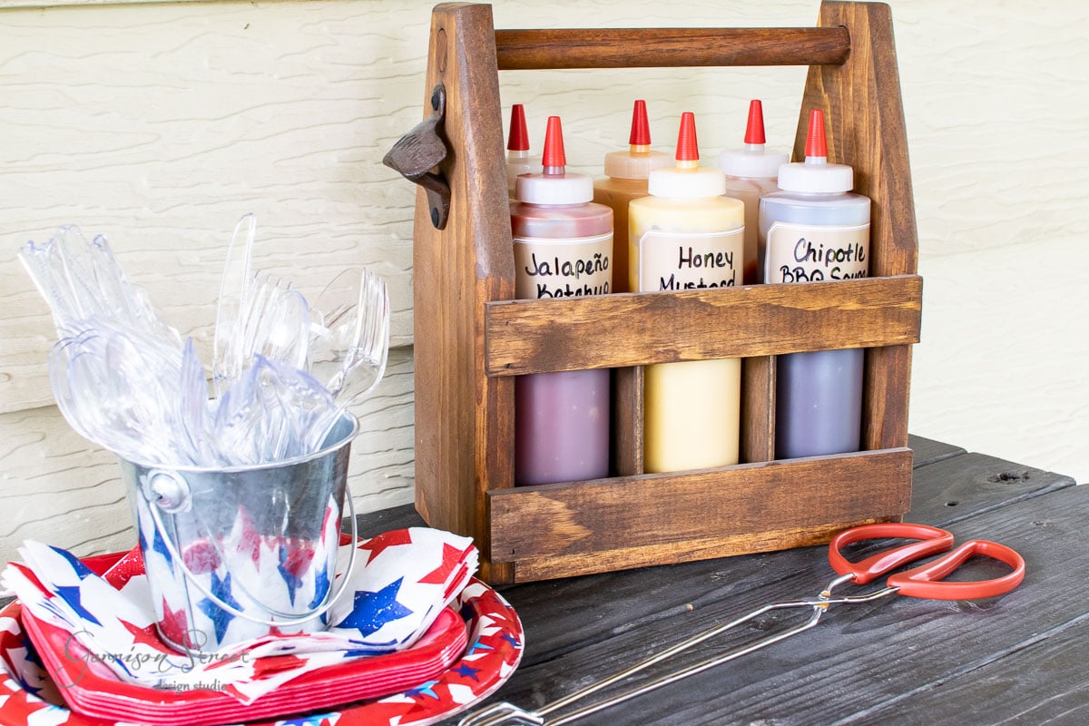 A wooden condiment caddy holds sauce bottles labeled Jalapeño Ketchup, Honey Mustard, and Chipotle BBQ Sauce. Nearby are star-patterned plates, a small metal bucket of plastic cutlery, and red-handled tongs on a dark wooden surface.