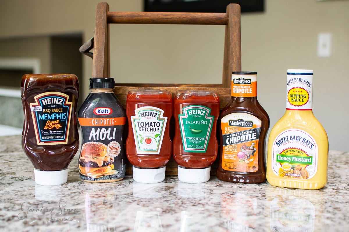A condiment caddy holds a selection of six bottles on a countertop, featuring Heinz Memphis BBQ Sauce, Kraft Chipotle Aioli, Heinz Tomato Ketchup, Heinz Jalapeño Ketchup, Sweet Baby Ray's Dipping Sauce, and Masterpiece Chipotle Marinade & Sauce.
