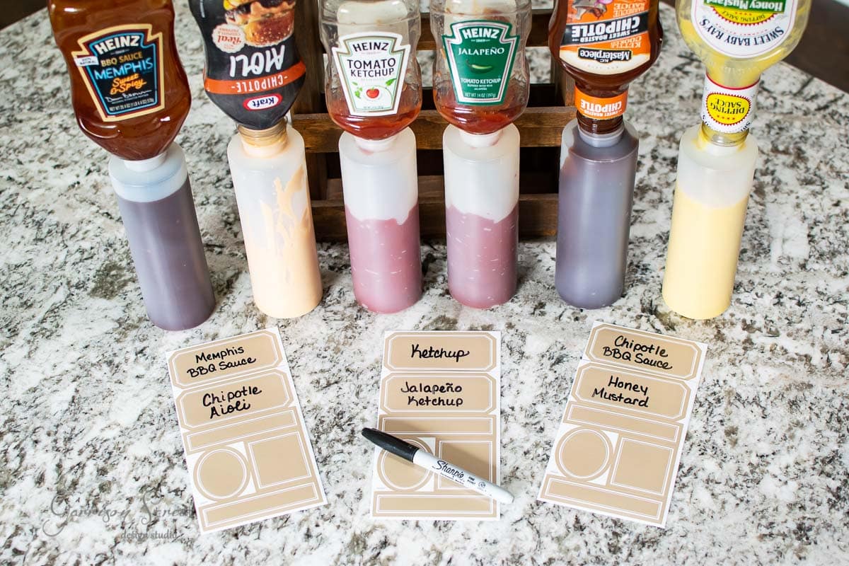Assorted condiment bottles are neatly displayed in a condiment caddy on the counter. Heinz sauces like BBQ, ketchup, and jalapeño feature labels resembling restaurant cue cards. A black marker rests beside them on the sleek marble surface.