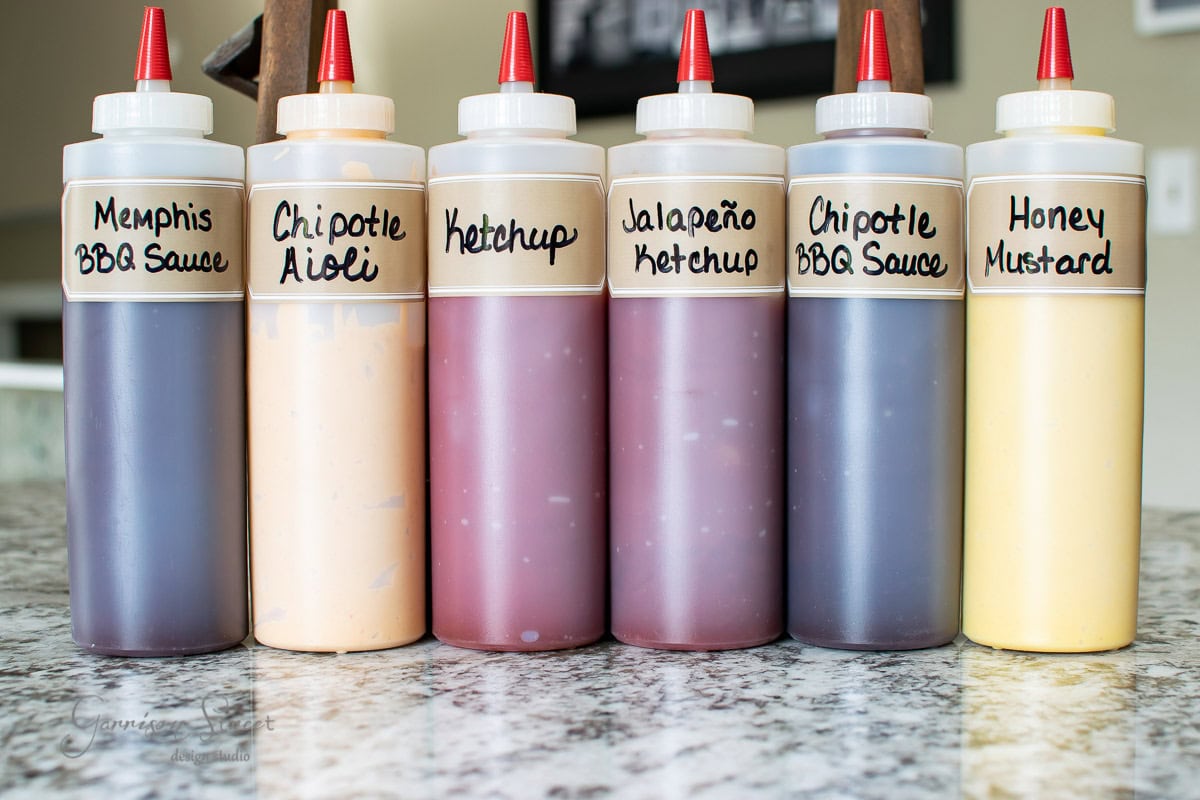 Six condiment squeeze bottles are neatly arranged in a condiment caddy on the marble countertop, each labeled: Memphis BBQ Sauce, Chipotle Aioli, Ketchup, Jalapeño Ketchup, Chipotle BBQ Sauce, and Honey Mustard.
