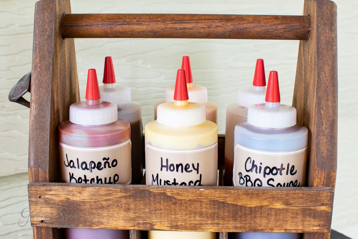 This condiment caddy elegantly holds eight squeeze bottles with vibrant red caps, labeled "Jalapeño Ketchup," "Honey Mustard," and "Chipotle BBQ Sauce." Neatly arranged in two rows, the bottles rest on a light-colored surface, ready to elevate any dining experience.