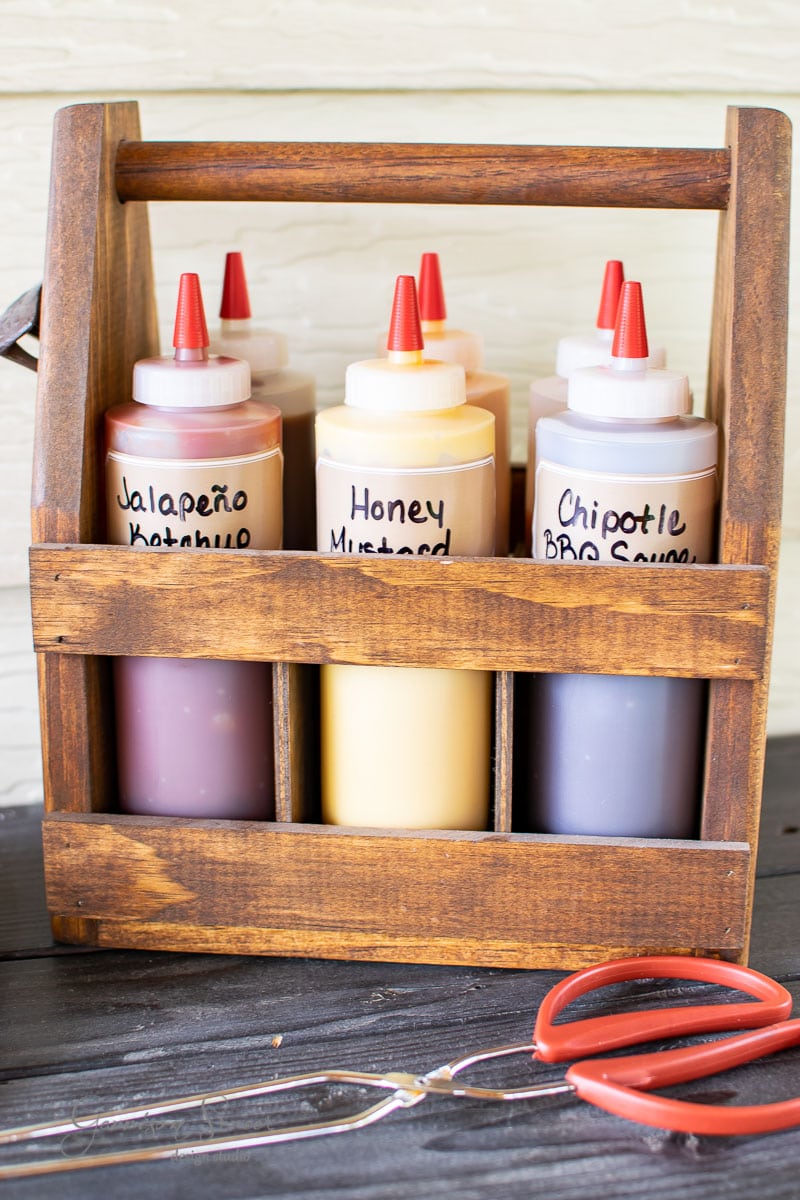 A wooden condiment caddy holds six squeeze bottles, each labeled with different sauces: Jalapeño Ketchup, Honey Mustard, and Chipotle BBQ Sauce. Nearby, tongs and scissors rest on the table for easy access.