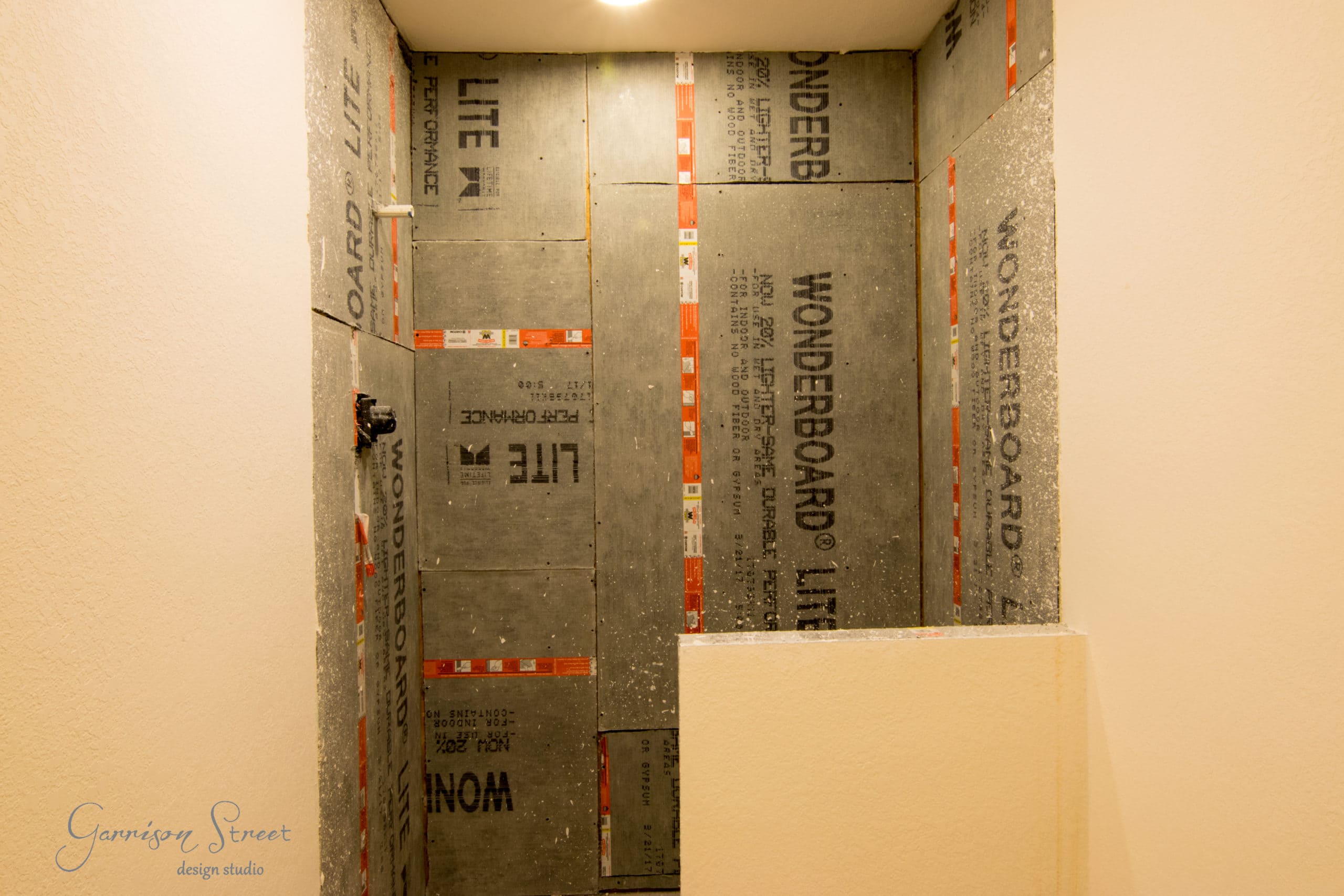 A shower area under construction features WonderBoard Lite cement backer boards on the walls. The space is illuminated by an overhead light. A partial wall is visible in the foreground, and plumbing fixtures are installed.