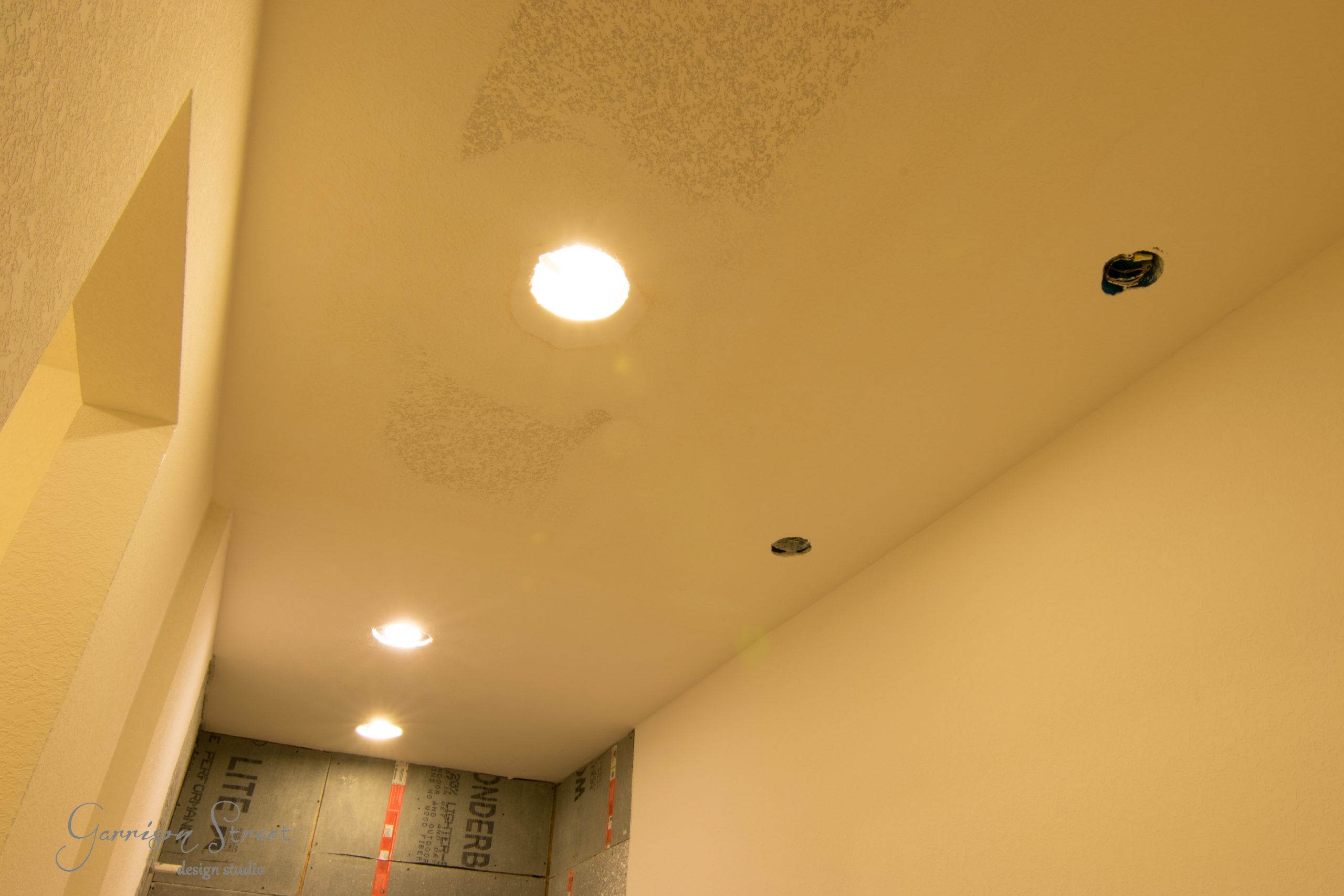 A room under renovation with three ceiling lights installed. The ceiling appears unfinished, with visible patches. The room has beige walls, and construction materials are partially visible on the right side.