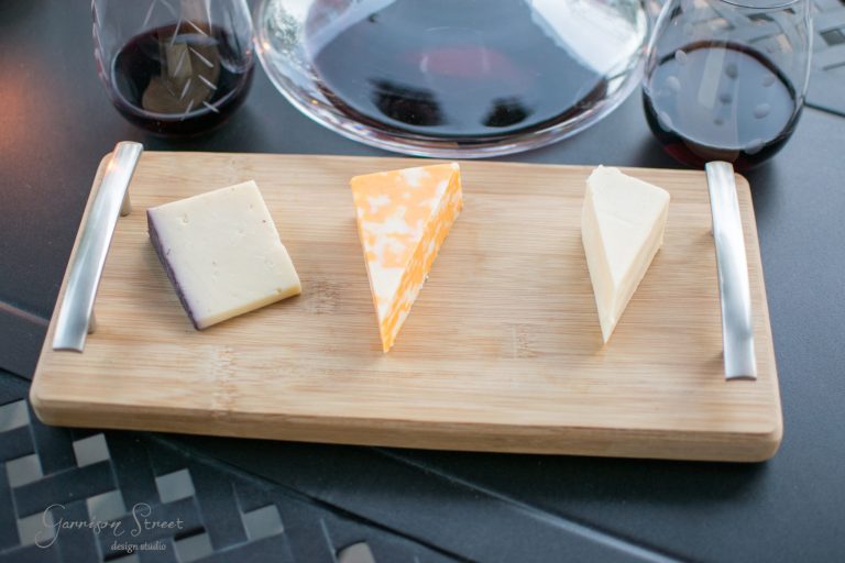 A cutting board with three types of cheese: a square light yellow piece, a marbled orange and white slice, and a triangular white wedge. Two glasses of red wine are in the background on a dark table.