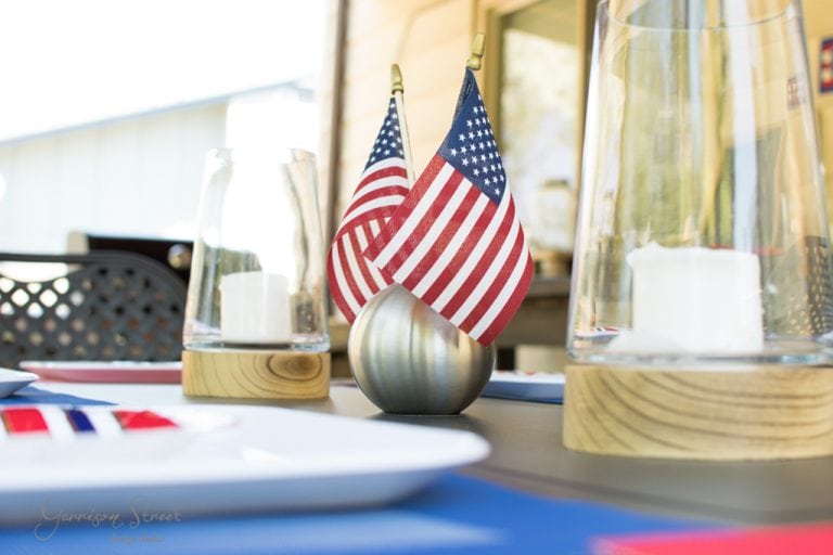 A table setting with two small American flags in a silver holder, surrounded by candle holders with white candles. The tableware includes plates and napkins, and the background shows partial views of outdoor furniture.