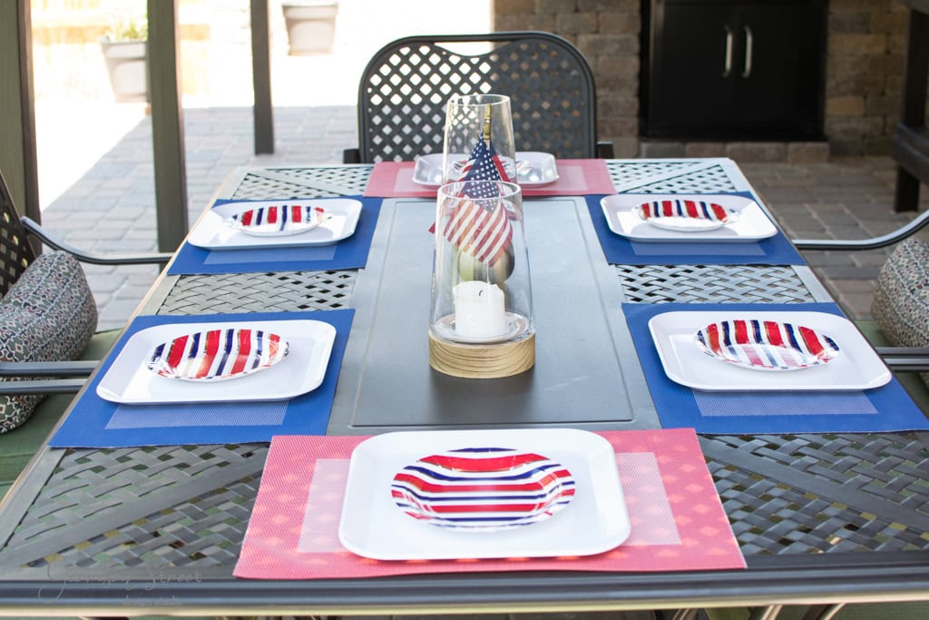 Outdoor dining table set for a patriotic occasion, featuring red, white, and blue striped plates on colorful placemats. A candle holder with a small American flag serves as a centerpiece. The setting is on a patio with a grill in the background.