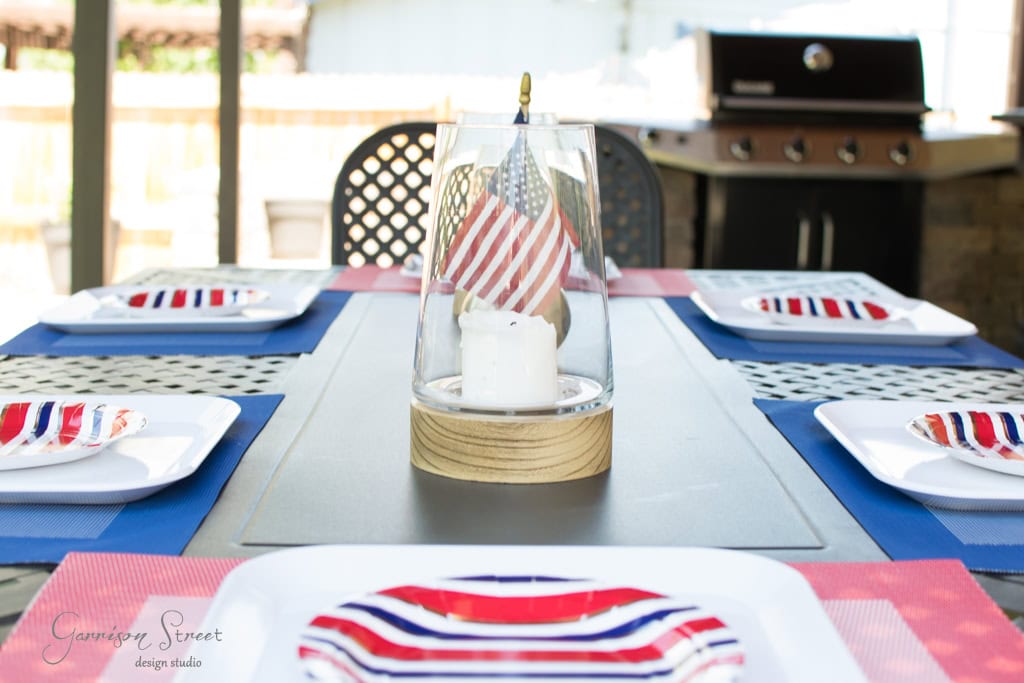 A table set for a meal with plates featuring red, white, and blue stripes. A centerpiece with an American flag and candle is in the middle. A grill is in the background.