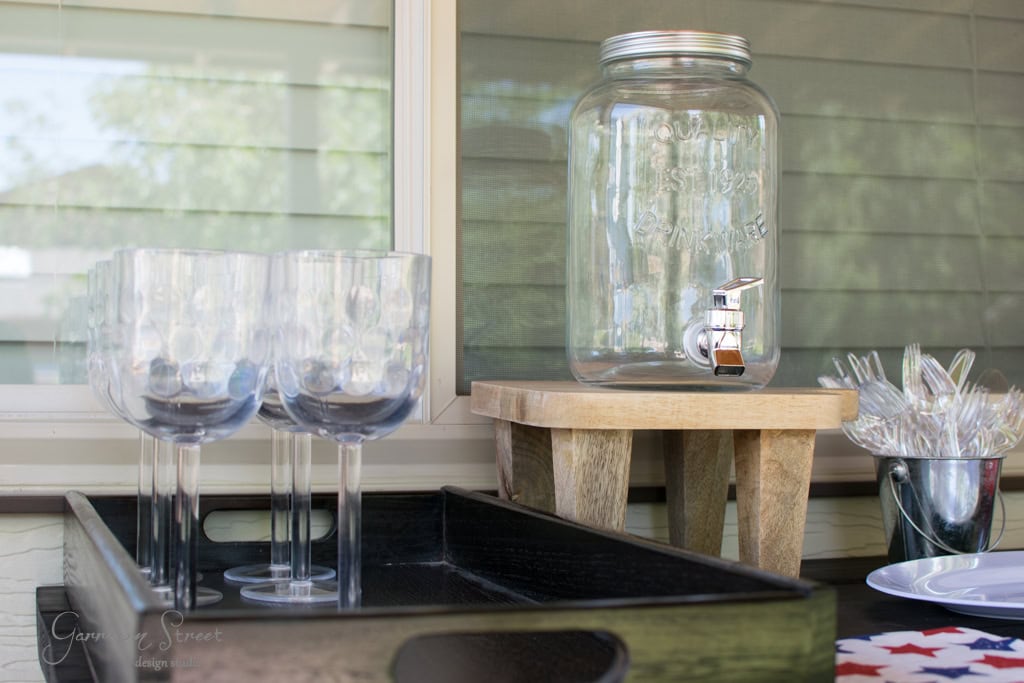 A clear glass beverage dispenser with a spigot is placed on a wooden stool. Next to it is a black tray holding several tall, transparent glasses and a metal container with plastic utensils on a table against a window.